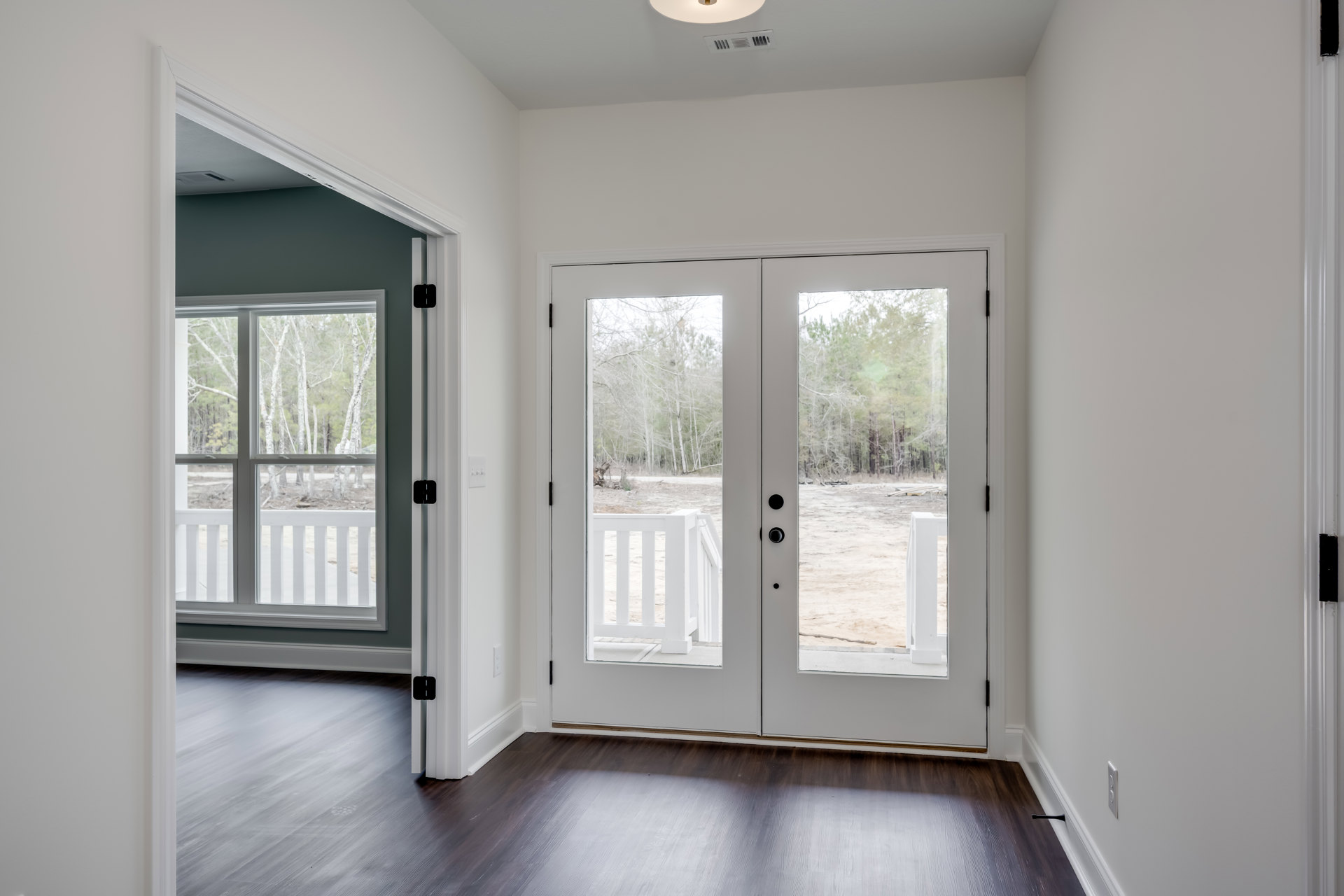 White double doors with glass panes, dark wood flooring, white porch railing, window showing trees outside, white walls and trim
