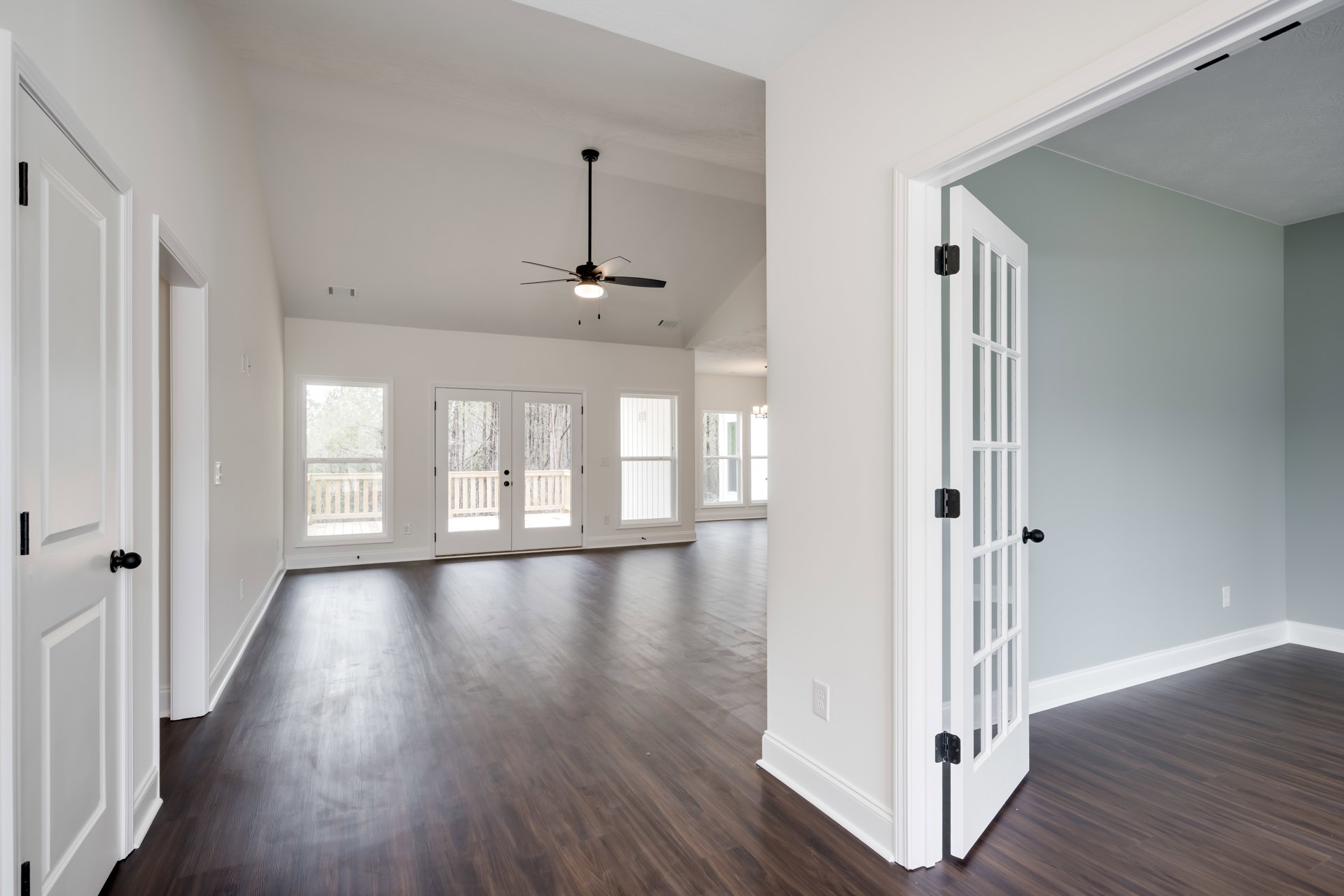 Spacious room with two ceiling fans, wood laminate flooring, double glass-panel doors, white door with black handle, large windows with blinds, view of deck and trees outside