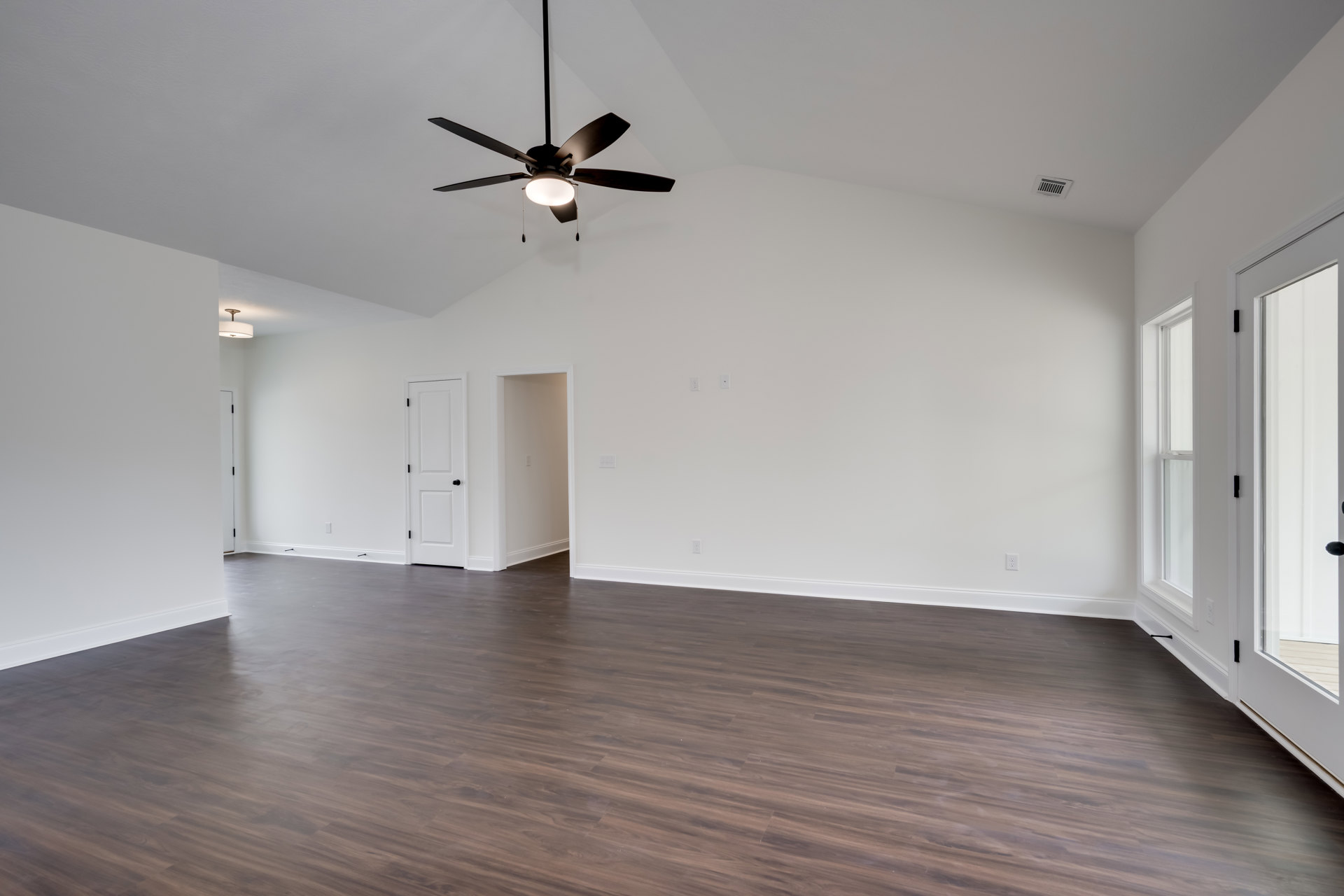 Spacious room with wood flooring, white walls, ceiling fan with light fixture, white door featuring black handle