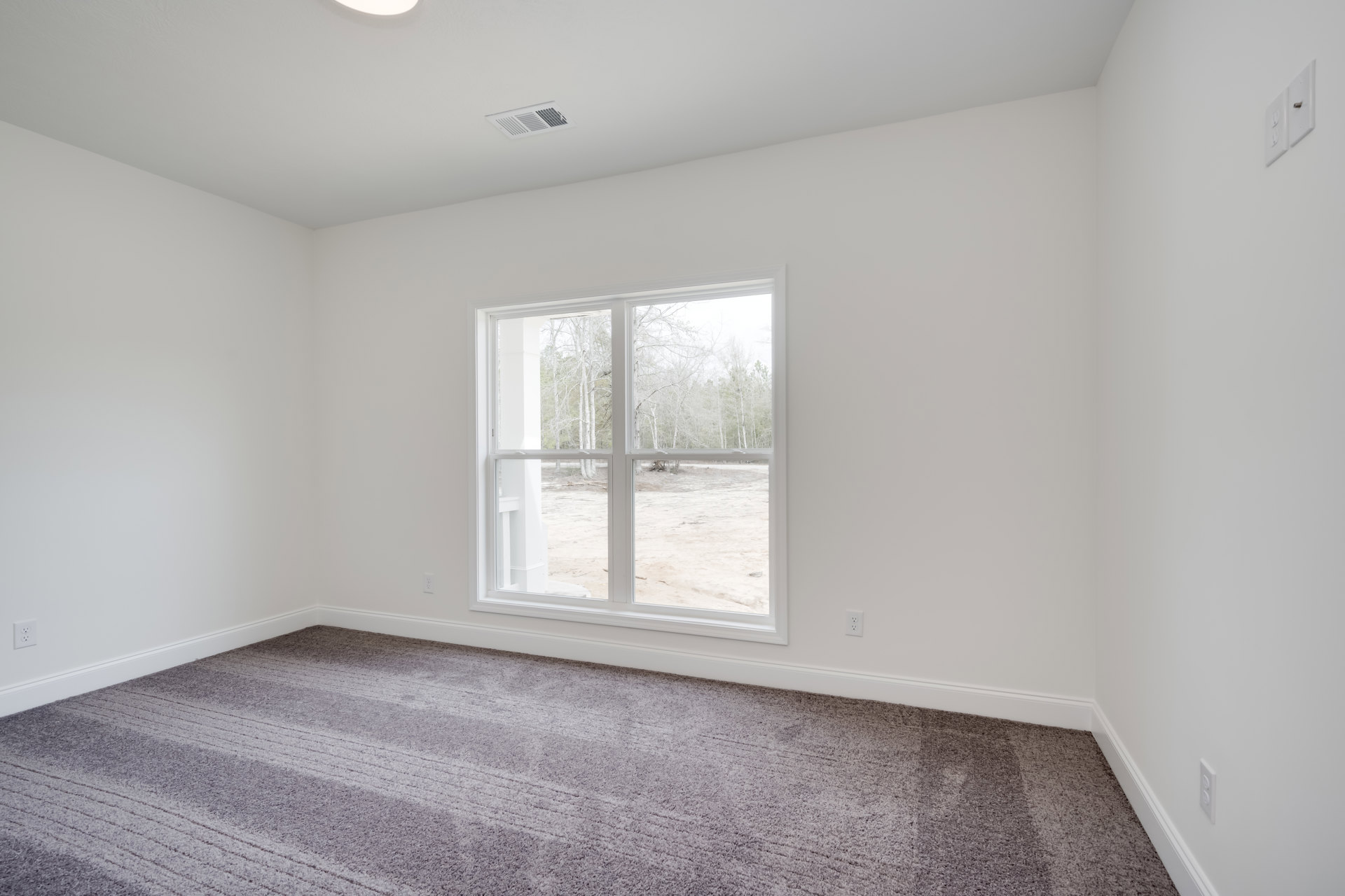 Carpeted room with a large window overlooking trees, white walls, ceiling vents, and a white electrical outlet with a silver fixture.