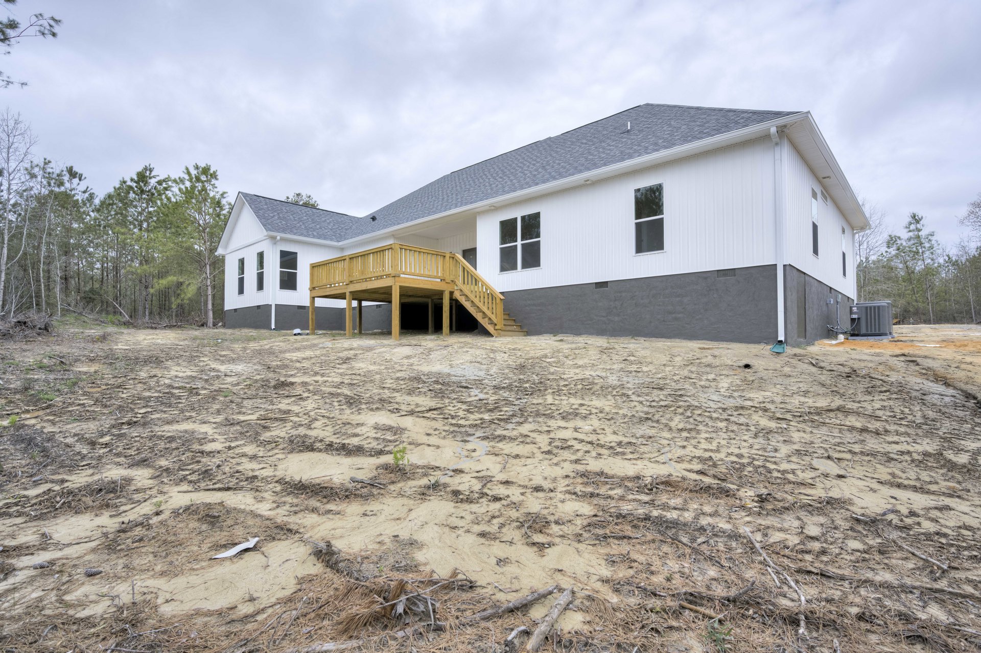 White house with wooden deck and railing, dirt yard with scattered sticks, large window reflecting trees, partly cloudy sky overhead