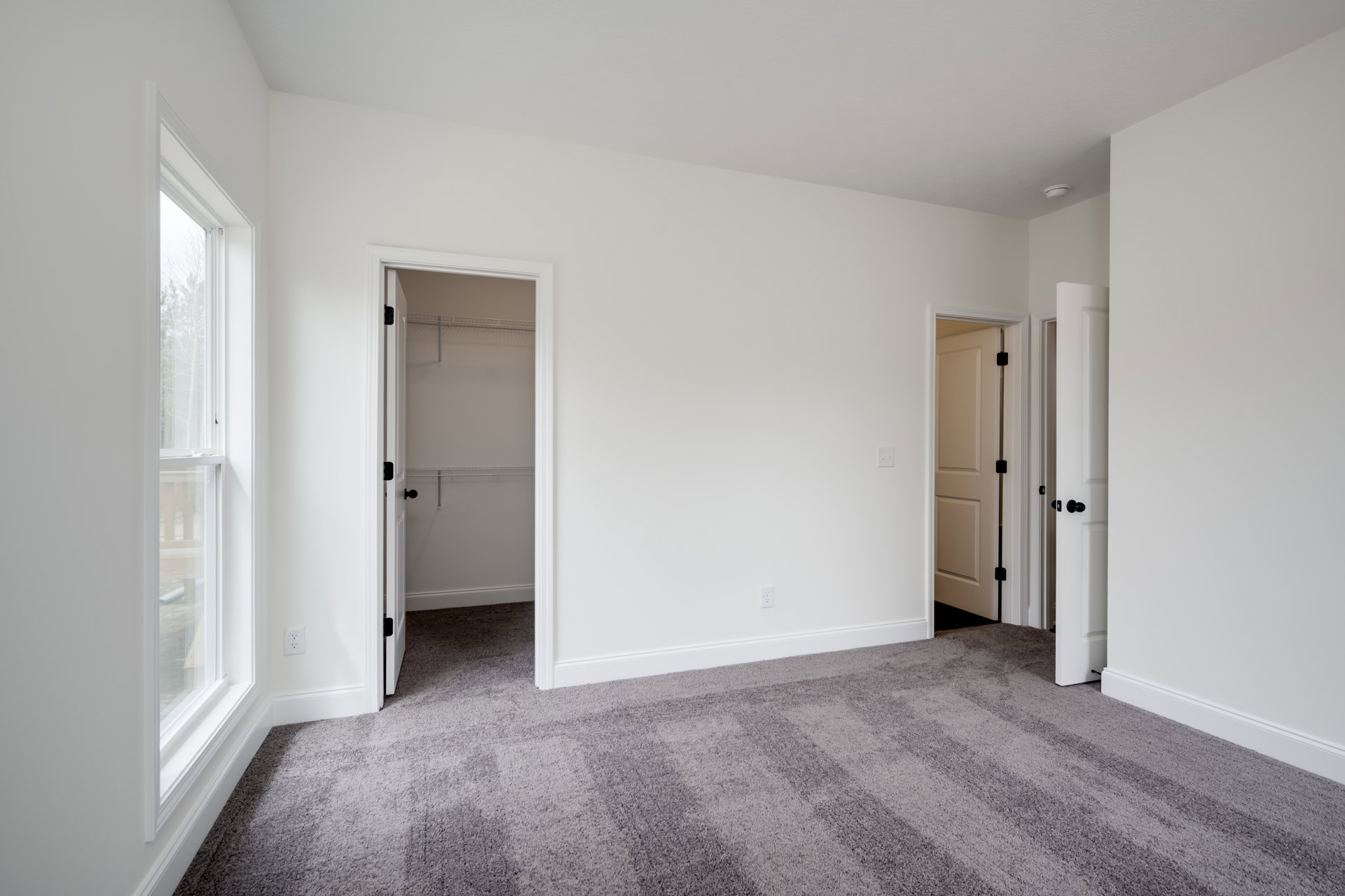 Bedroom with white walls, light carpet flooring, open doors, metal closet shelf, and a black sign on one door; roll of paper mounted on wall.
