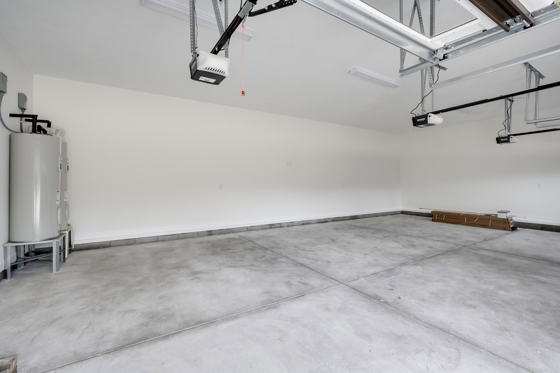 Spacious garage with white plaster walls, grey tile flooring with white trim, mechanical arm mounted on white ceiling, brown box with white tape, large white tank with warning
