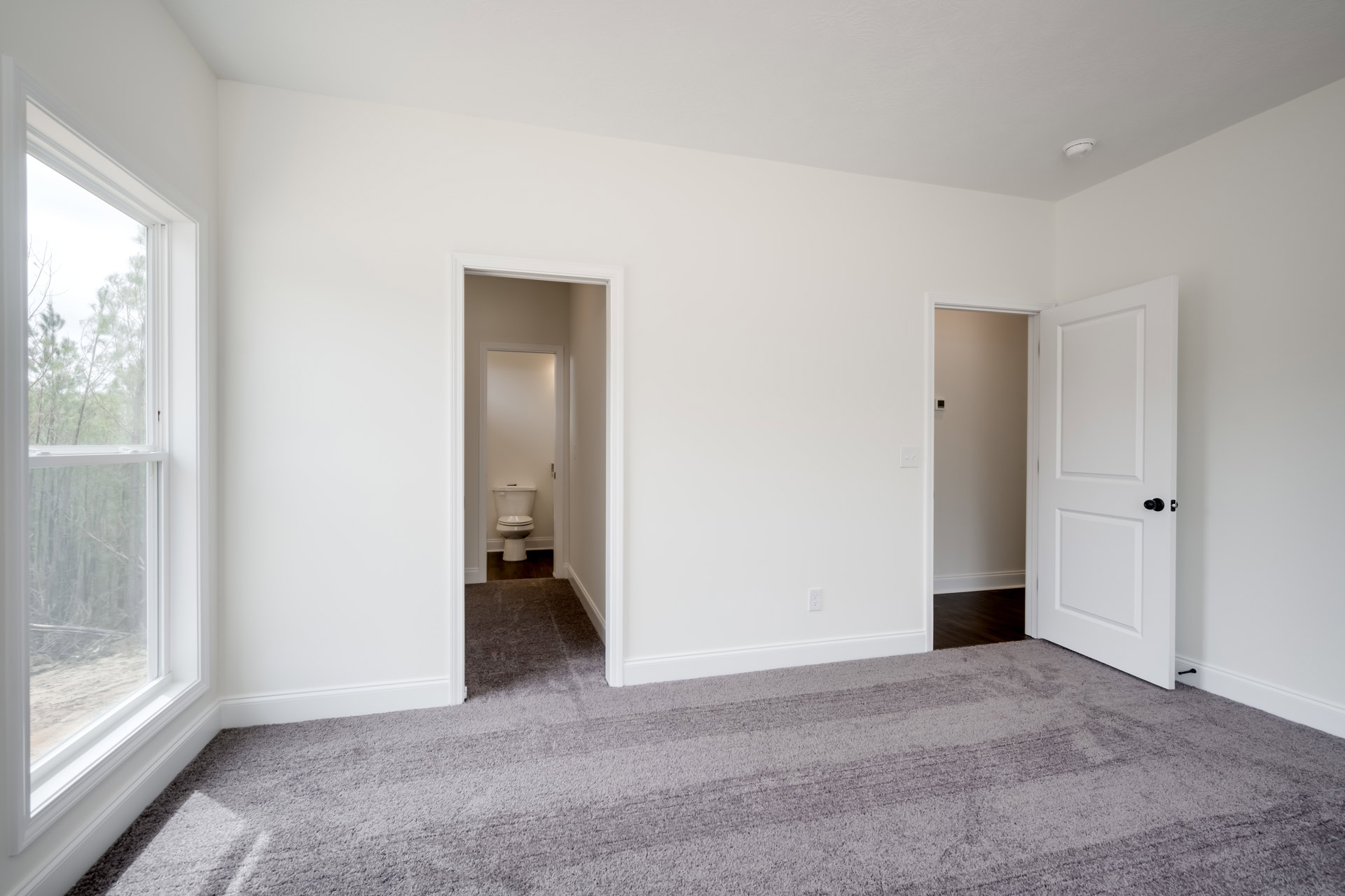 Bathroom with white toilet, open white door featuring black knob, carpeted flooring, window overlooking trees, plaster walls, and laminate ceiling