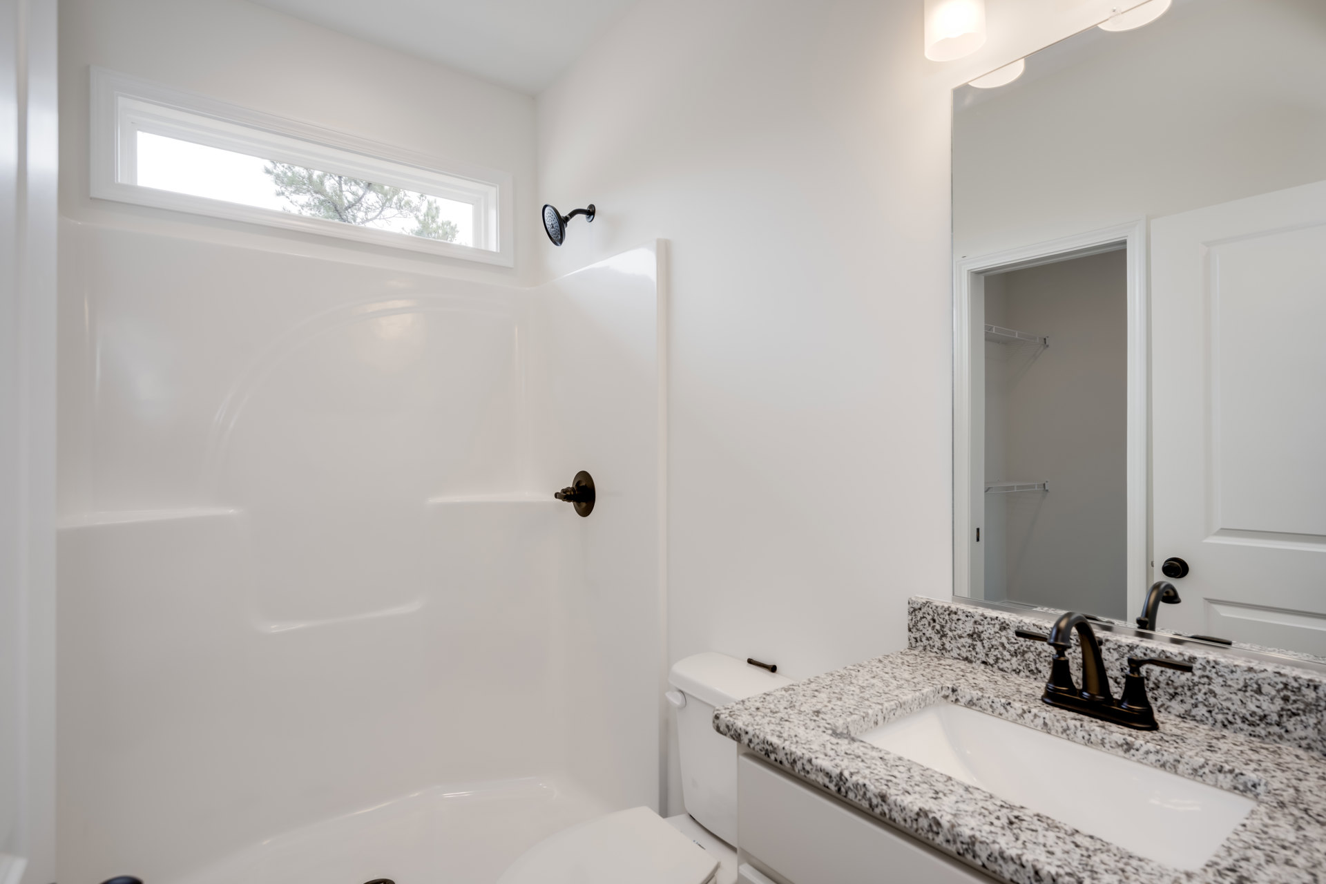 Modern bathroom with white tile walls, black faucet on rectangular sink, wall-mounted shower head, window above tub, and white toilet