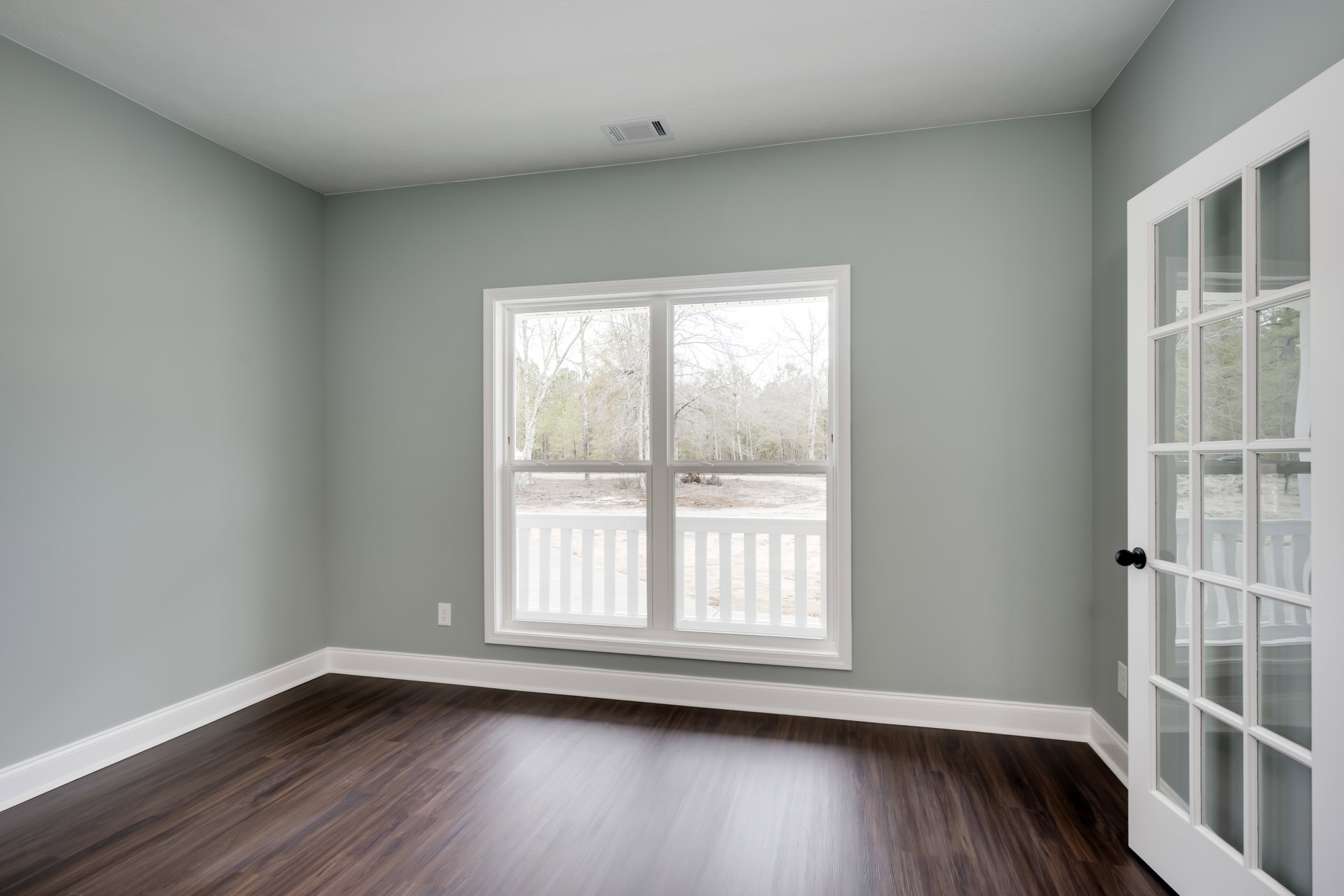 Hardwood floor room with white walls, large window overlooking porch, glass-paneled door, white window sill, ceiling vent