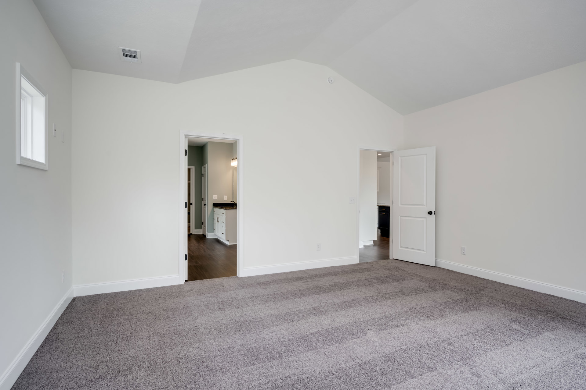 White-walled room with light carpet flooring, open white door with black knob, rectangular window, and white kitchen cabinet with drawers