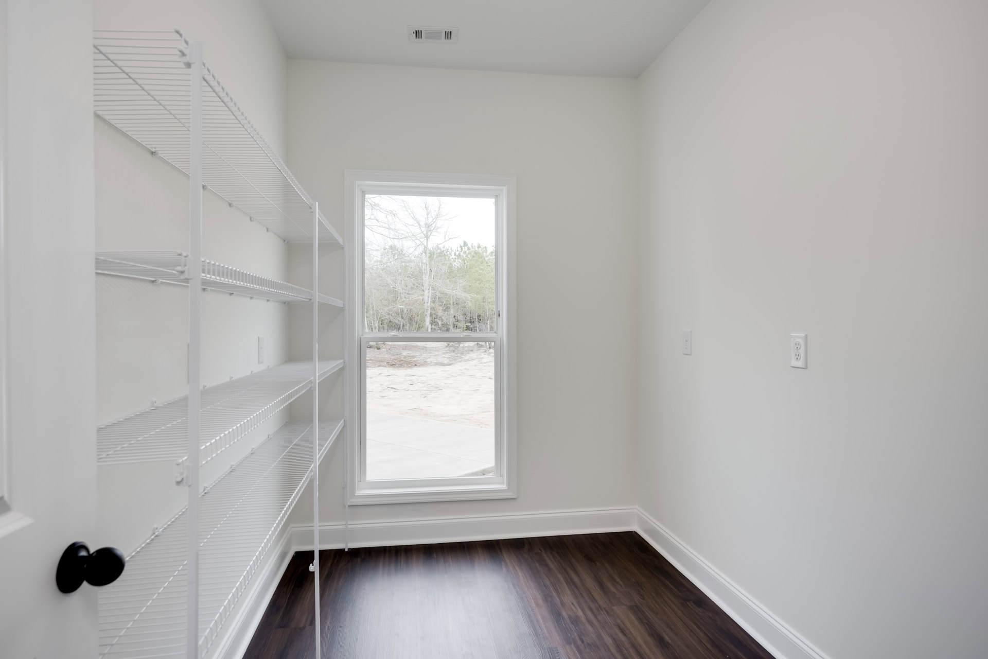 White built-in shelves along a plaster wall, dark laminate wood flooring, large window with view of green trees, ceiling with recessed lighting.