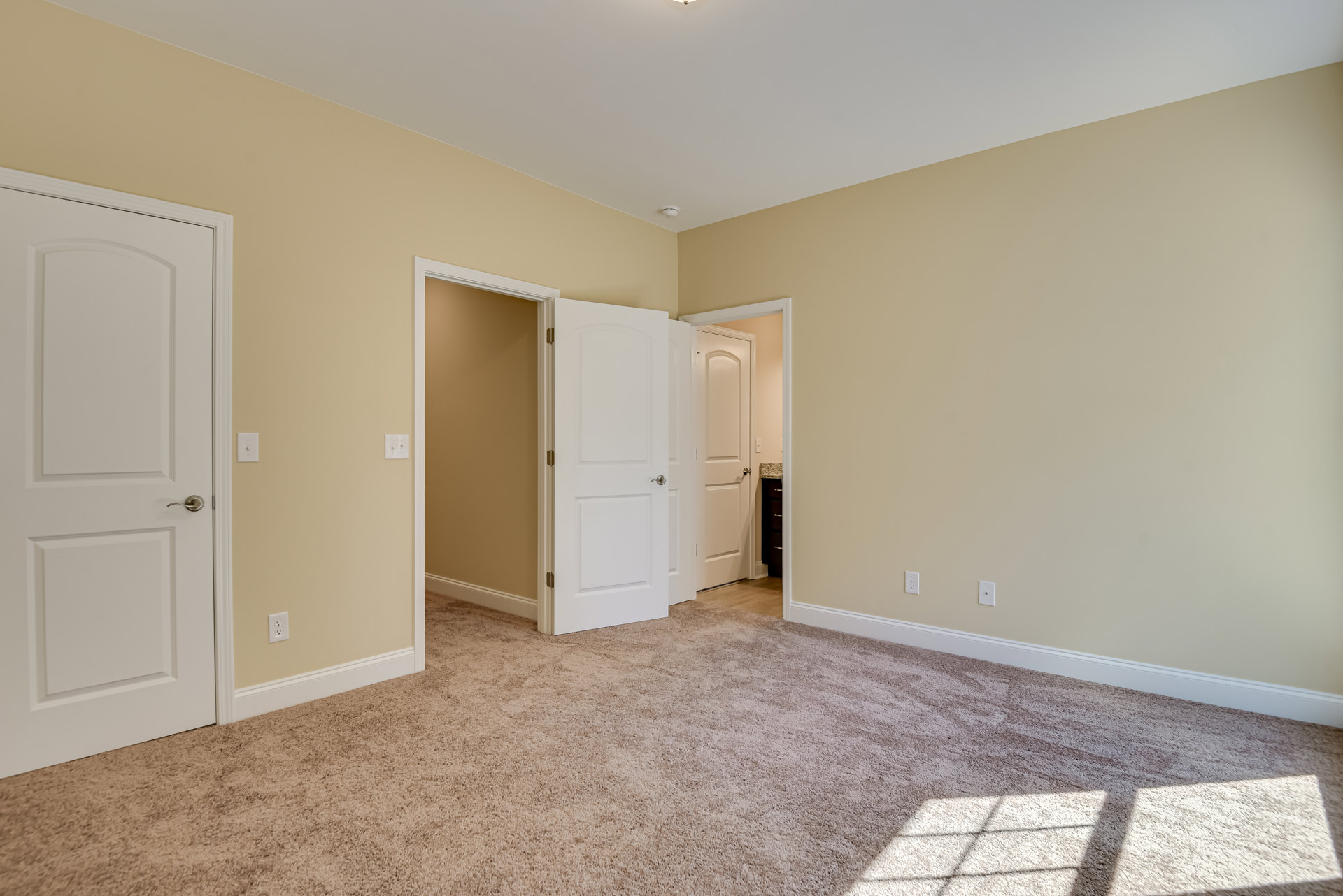 Beige walls, white paneled doors with silver handles, carpeted floor featuring a star pattern, white door partially open, wall-mounted light fixture adjacent to door, simple