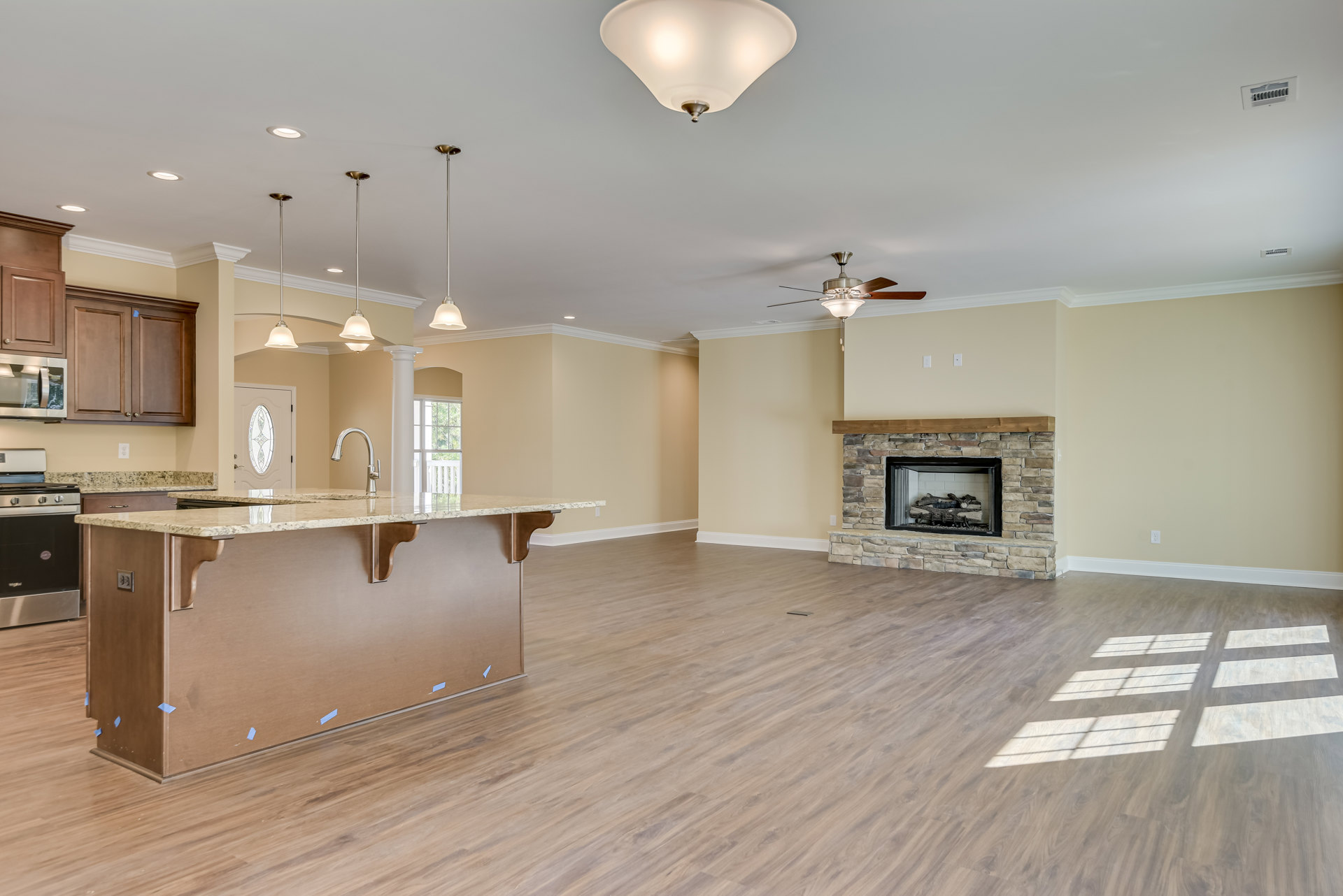 Living room featuring wood flooring, stone fireplace with stacked logs, kitchen island with light-colored countertop, built-in bar cabinetry, and modern lighting fixtures.