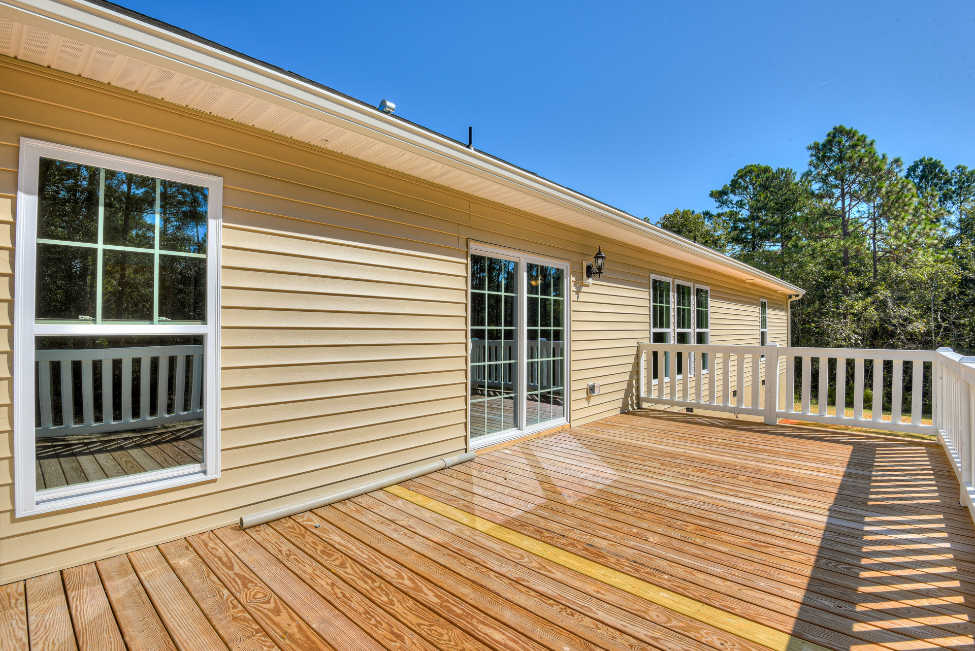 Wood deck with white railing, glass door with white frame, windows with white trim, light-colored siding, trees and sky in background