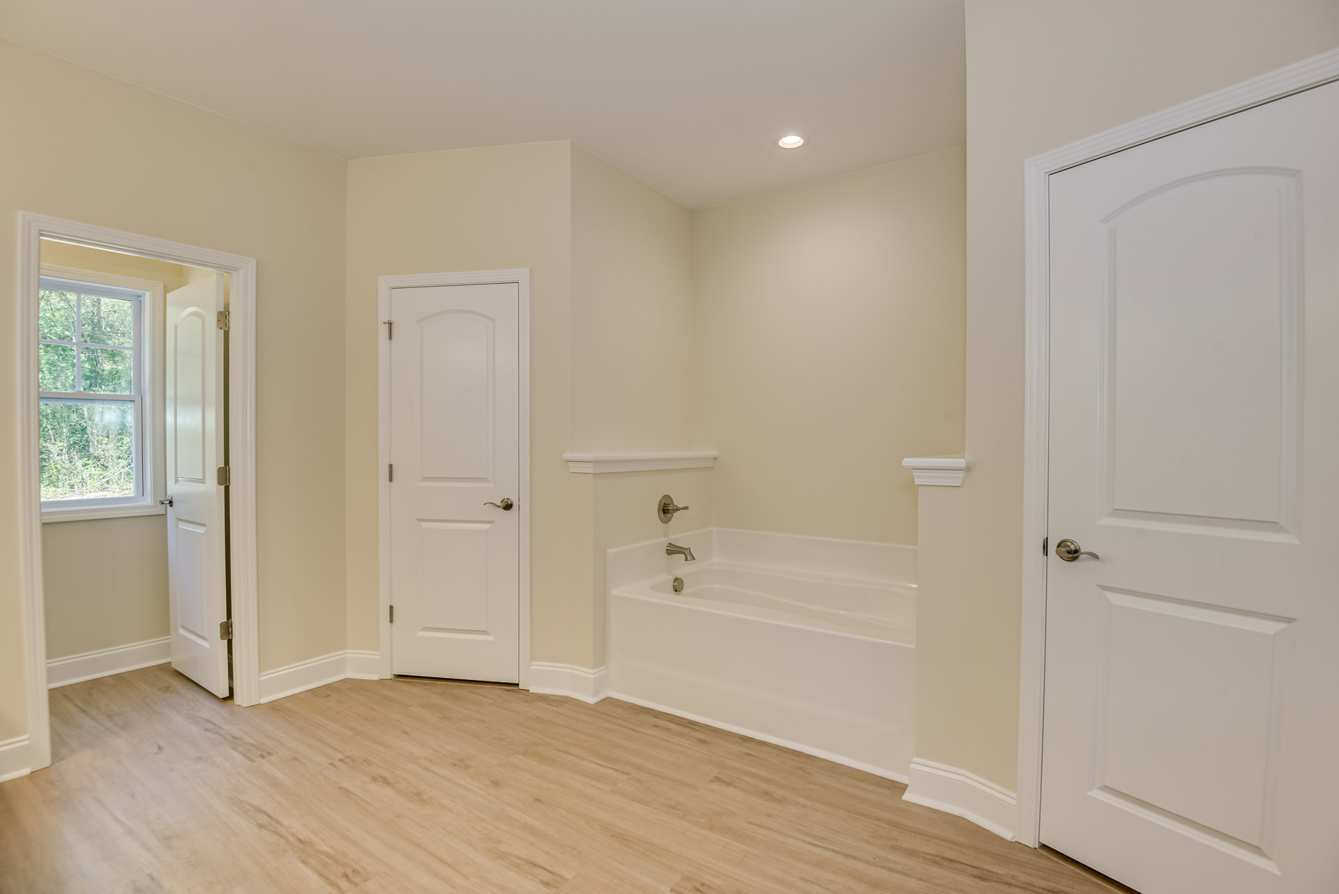 Bathroom with white walls, freestanding bathtub, wood flooring with white trim, white door with silver handle, window showing trees outside