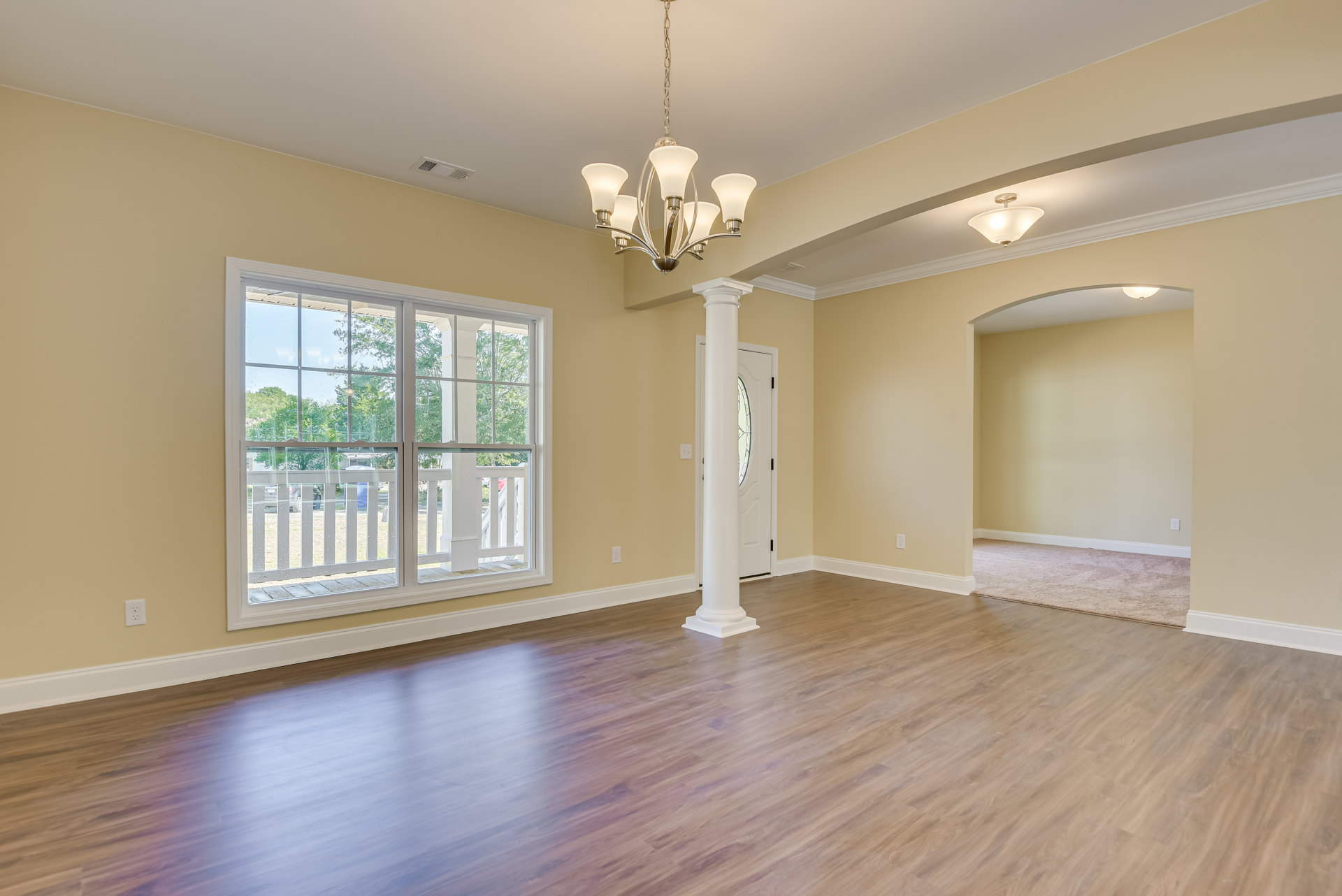 Wood flooring and white column in a spacious room, chandelier hanging from ceiling, large window with white railing, neutral walls with molding, carpet partially visible.