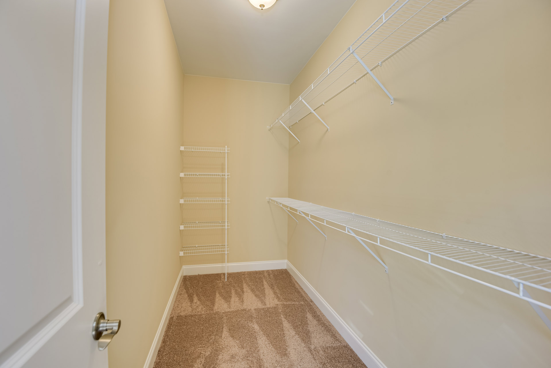 Walk-in closet with white wire shelving, beige carpet flooring, and a white door handle visible in the foreground