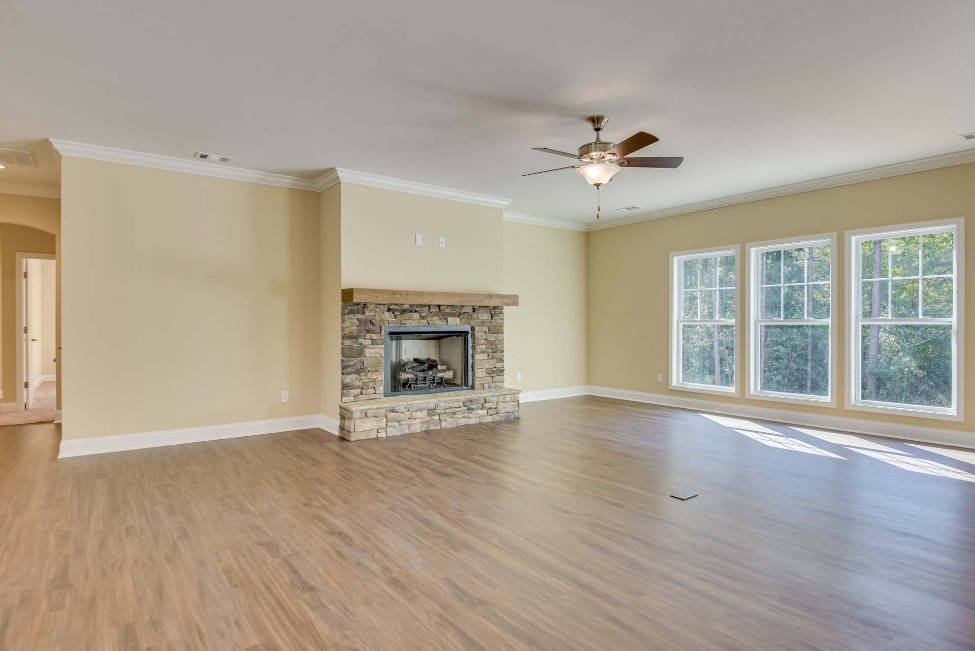 Living room with hardwood floor, central stone fireplace with stacked logs, large window showing trees outside, ceiling fan with light fixture above