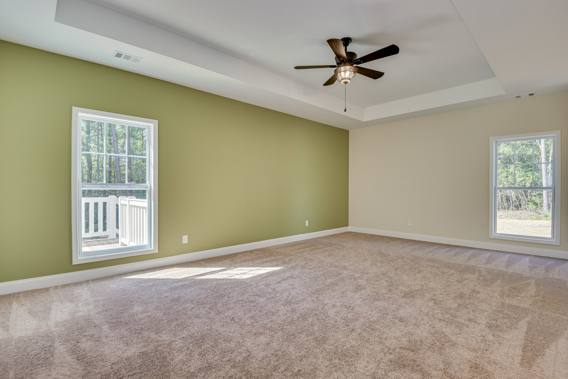 Carpeted bedroom with green accent wall, ceiling fan with light fixture, large window overlooking trees, white trim and molding