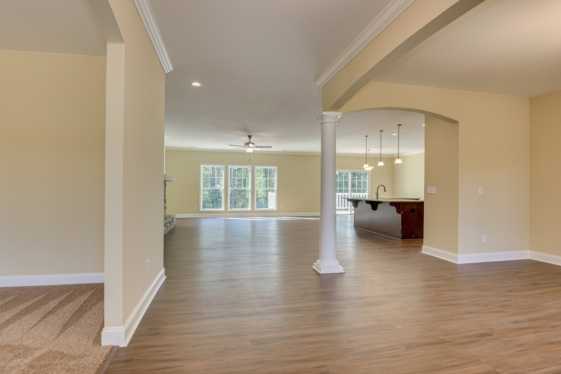 Spacious open floor plan featuring white columns, wood flooring, row of windows with tree views, white pillar, countertop with faucet, and ceiling fan with light fixture