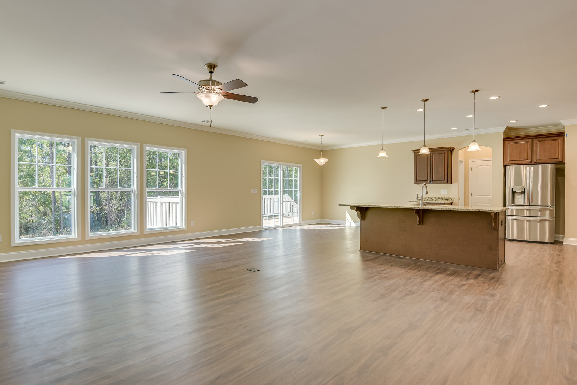 Spacious room featuring wood flooring, a bar area with stainless steel refrigerator and countertop, ceiling fan, white-framed windows overlooking trees, and plaster walls.