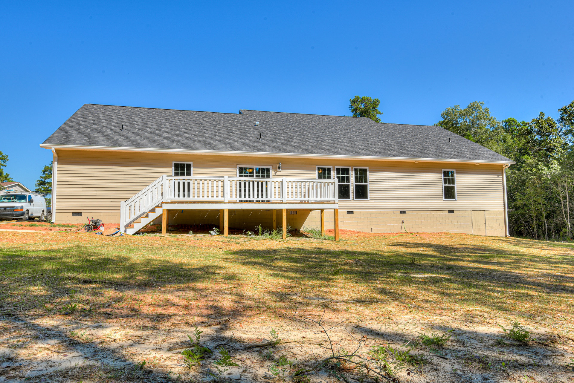 Two-story house with light siding, elevated deck featuring white railing, grassy yard bordered by trees, and a white van parked along the street.