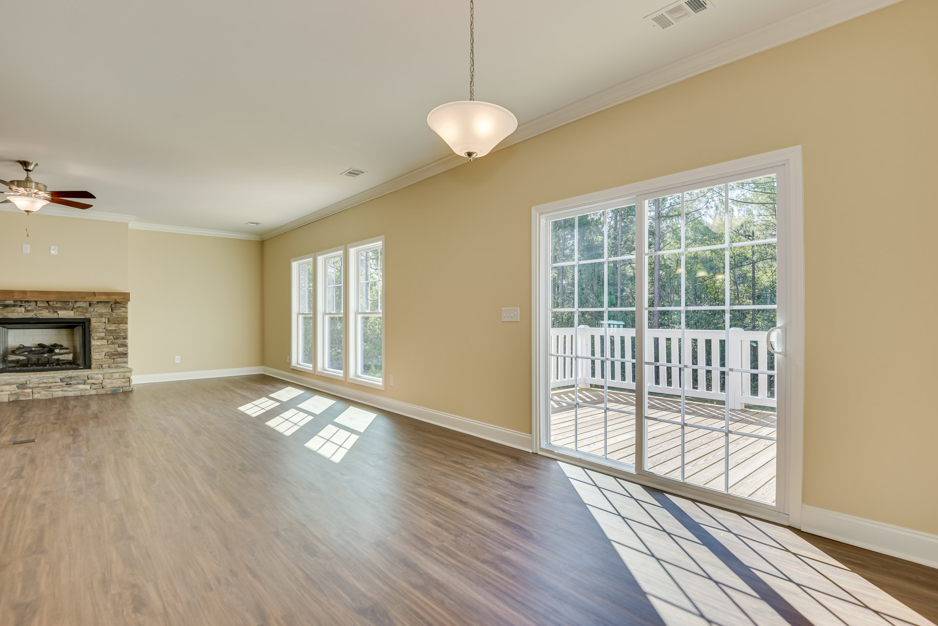 Open living room with wood flooring, white walls, ceiling fan light fixture, sliding glass doors leading to a deck overlooking trees