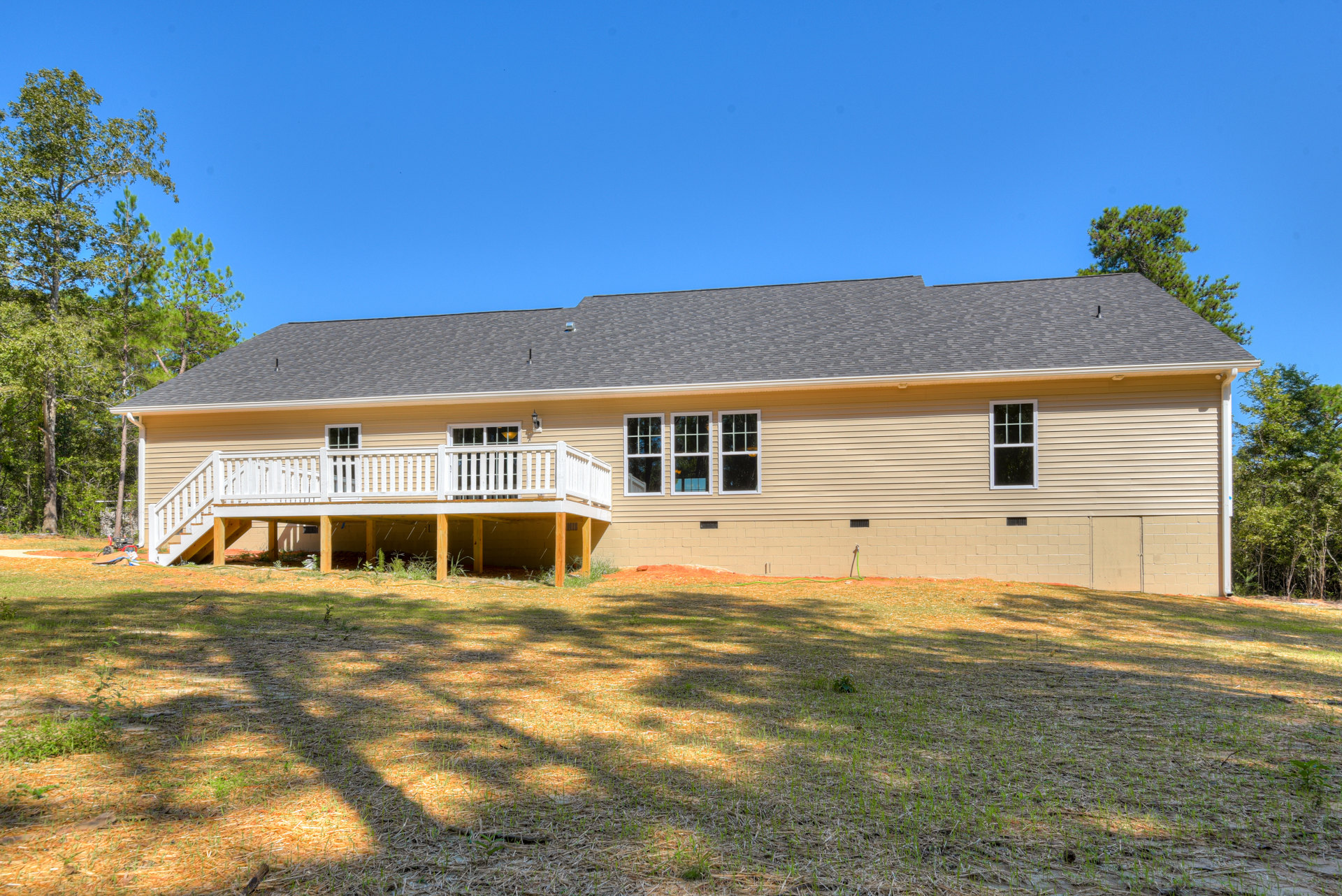 Two-story house with gray siding, white-trimmed windows, elevated wooden deck with white railing, covered porch, and manicured grass yard under clear blue sky
