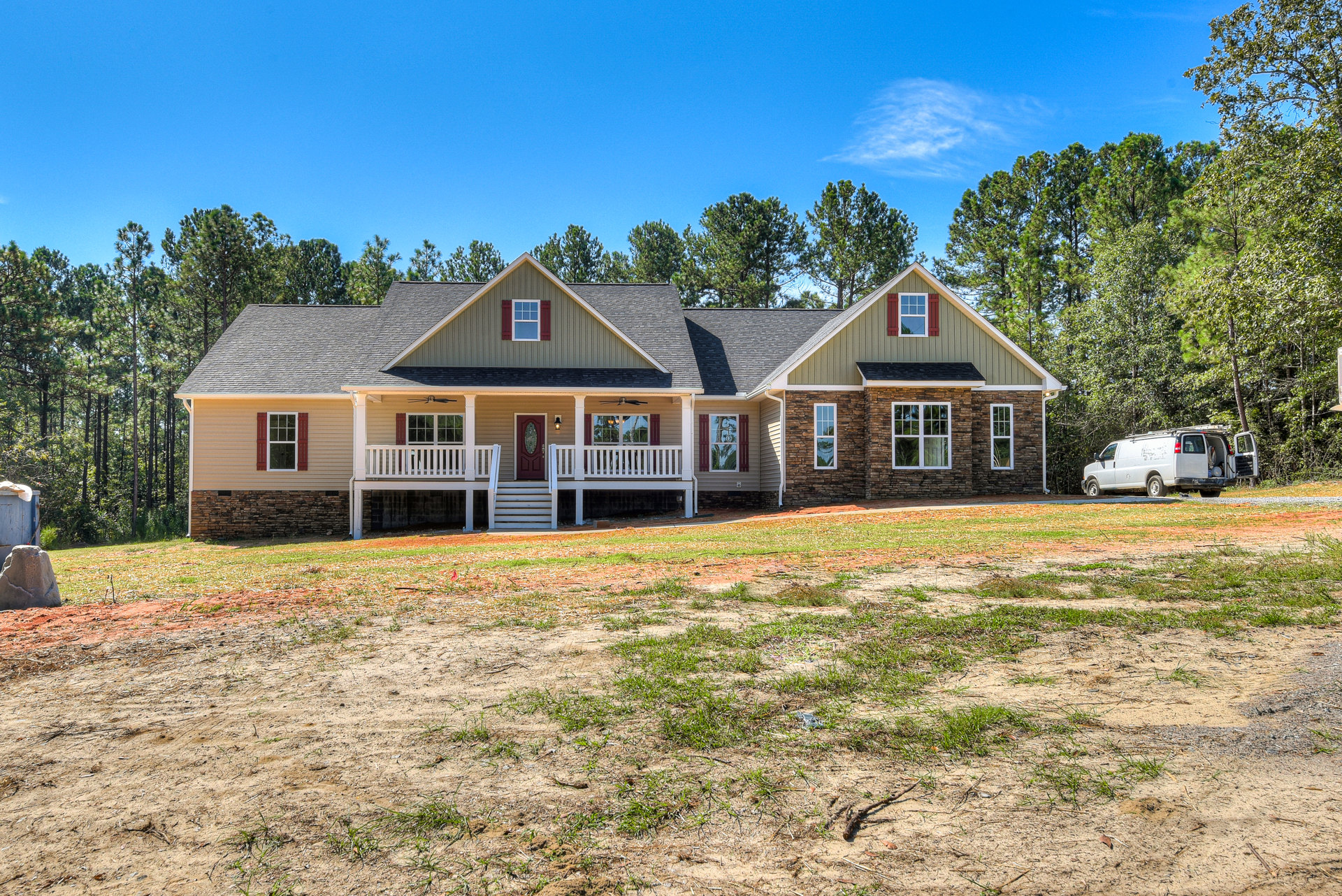 Brick house with white porch railing, large grassy yard, white van parked beside, multiple windows, mature trees in background