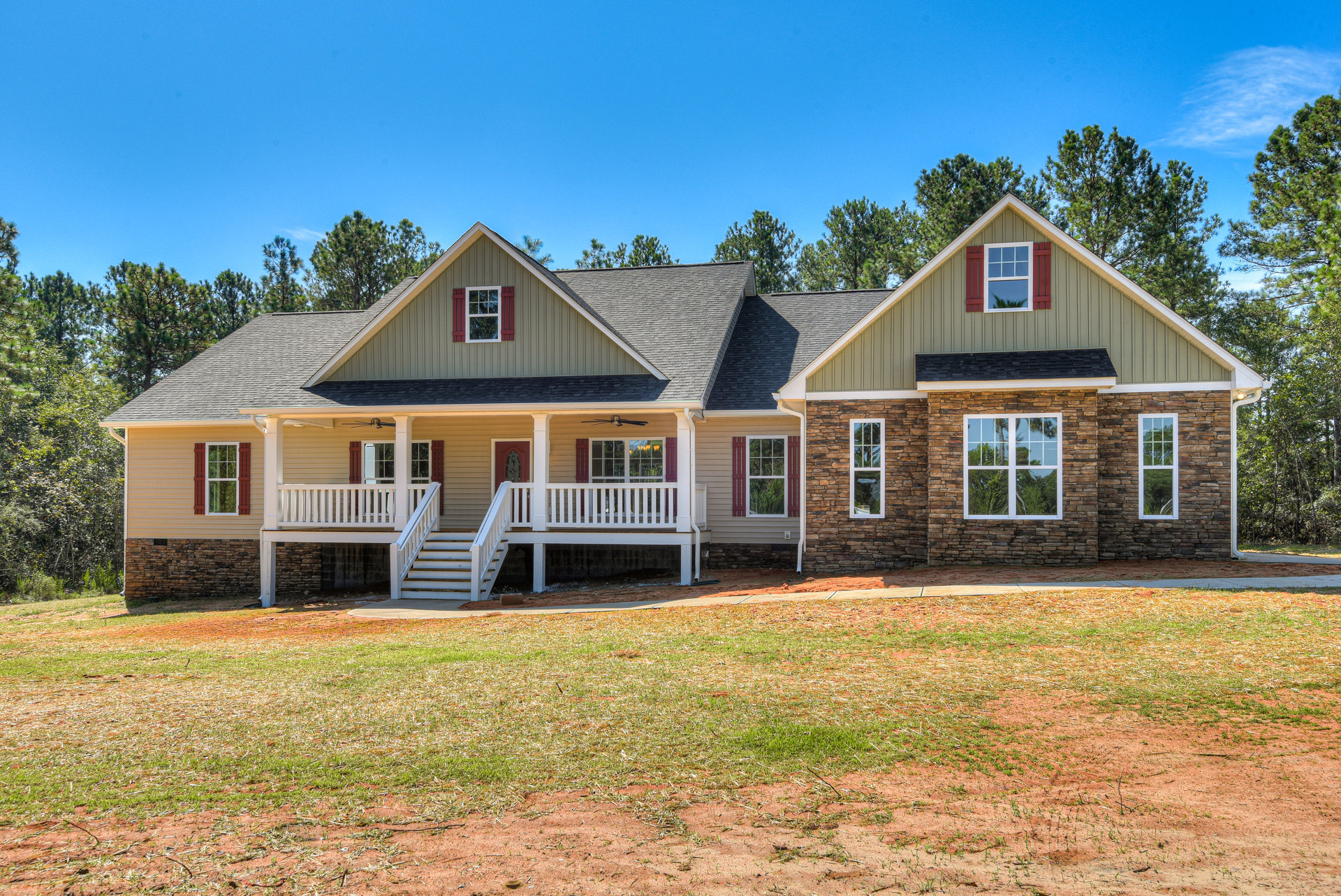 Two-story house with stone porch, white railings, and large windows, surrounded by green lawn and mature trees under a clear blue sky