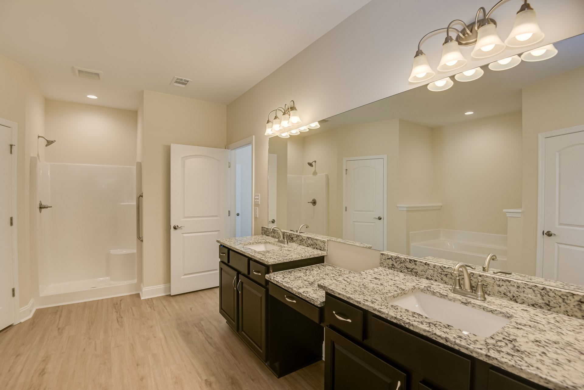 Double vanity with undermount sinks, quartz countertop, large framed mirror, row of chrome light fixtures, white shaker cabinets, tile flooring, white door with brushed nickel