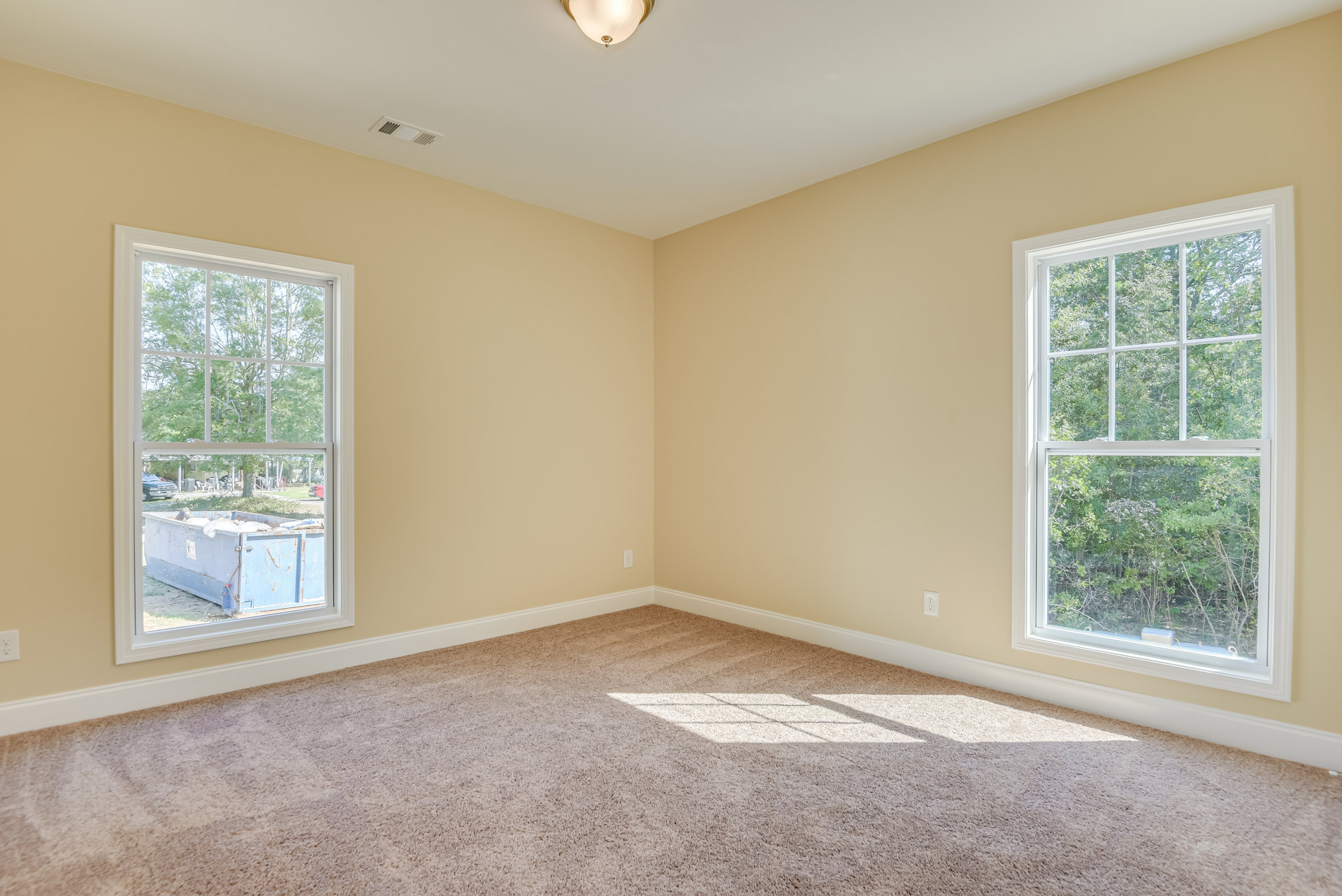 Beige-walled room with carpeted floor, two windows showing trees and a blue dumpster outside, sunlight streaming onto carpet