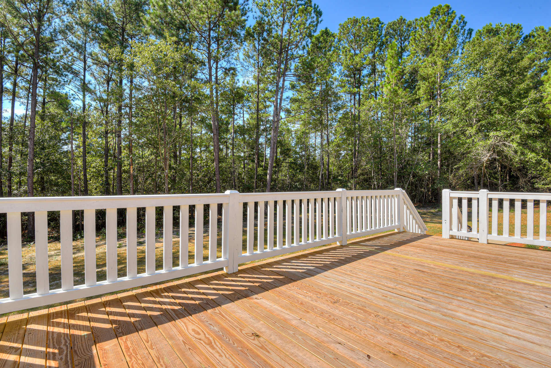 Wooden deck with white railing overlooking grassy yard and tall trees in the background