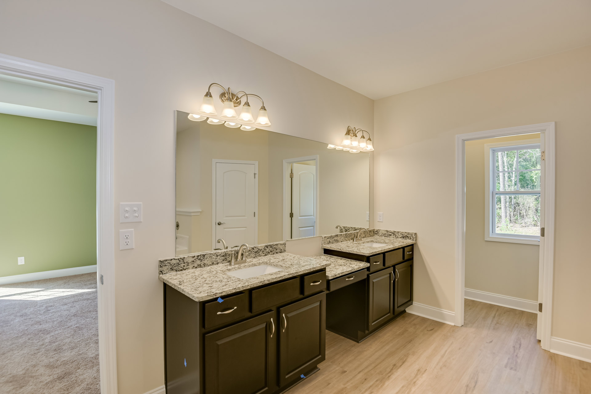 Bathroom with wide framed mirror above double white sinks, stone countertop, modern cabinetry, silver faucets, window showing trees, white door with silver handle, recessed