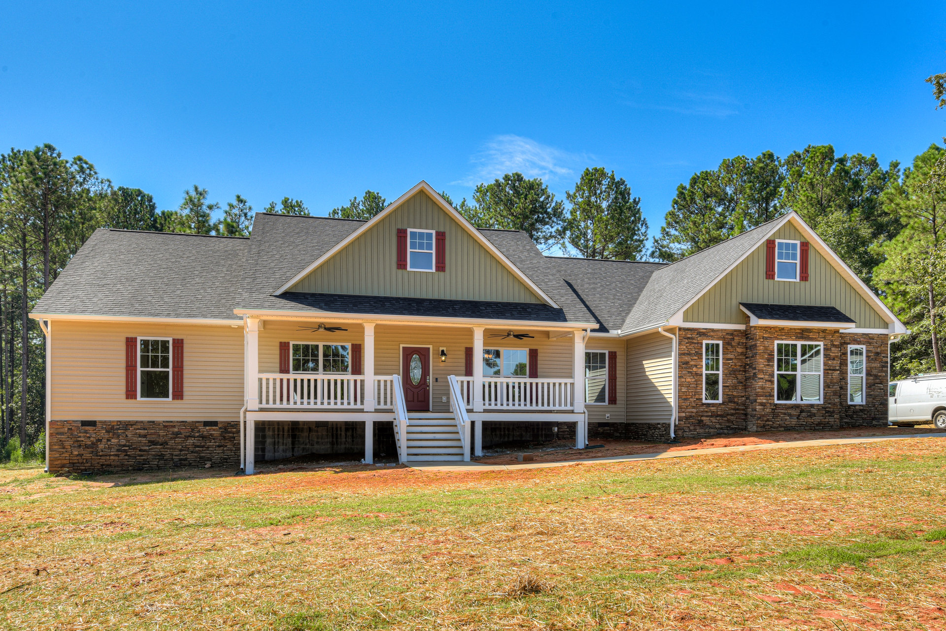 Two-story house with wide front porch, red front door, white stairs, red window shutters, grass lawn, and white van parked in driveway