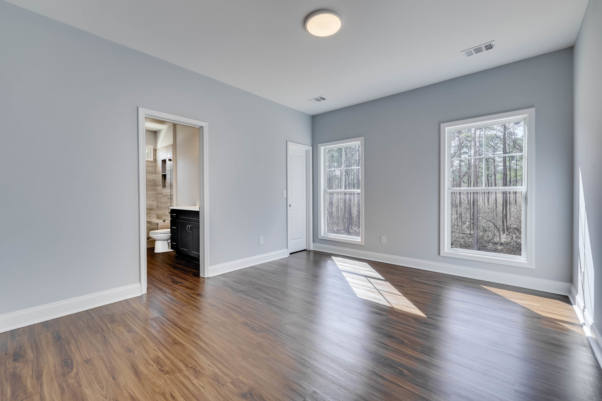 Wood flooring in a room with a black cabinet, white toilet, ceiling light fixture, and a door; window reveals trees outside.