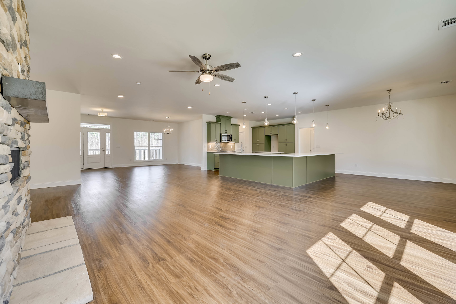 Open-concept kitchen with white island, wood flooring, green accent wall, ceiling fan with light, and chandelier hanging from plaster ceiling.