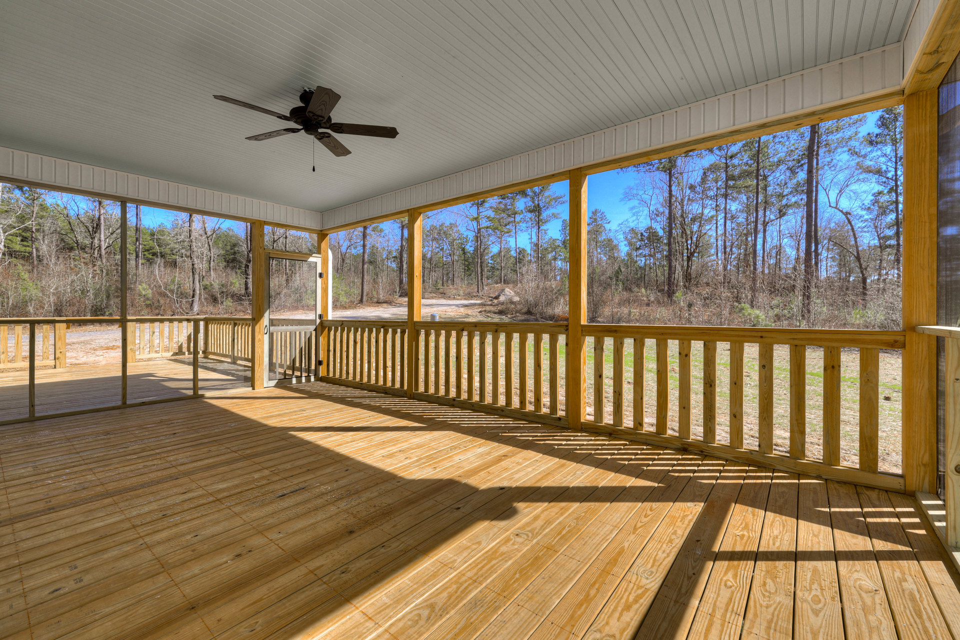 Wooden deck with railing, ceiling fan with wood blades overhead, surrounded by trees and grassy yard in the background
