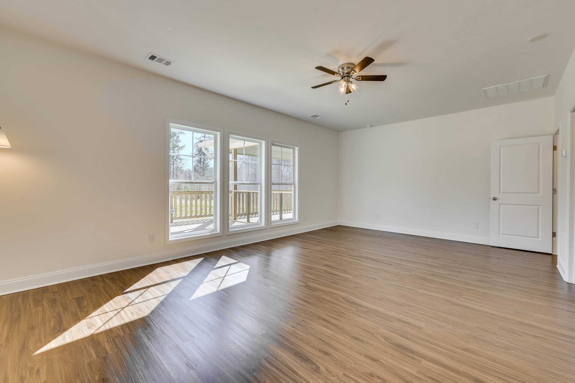 Ceiling fan with lights above hardwood floor, white door with silver knob, window with wood railing overlooking trees, floor lamp near wall