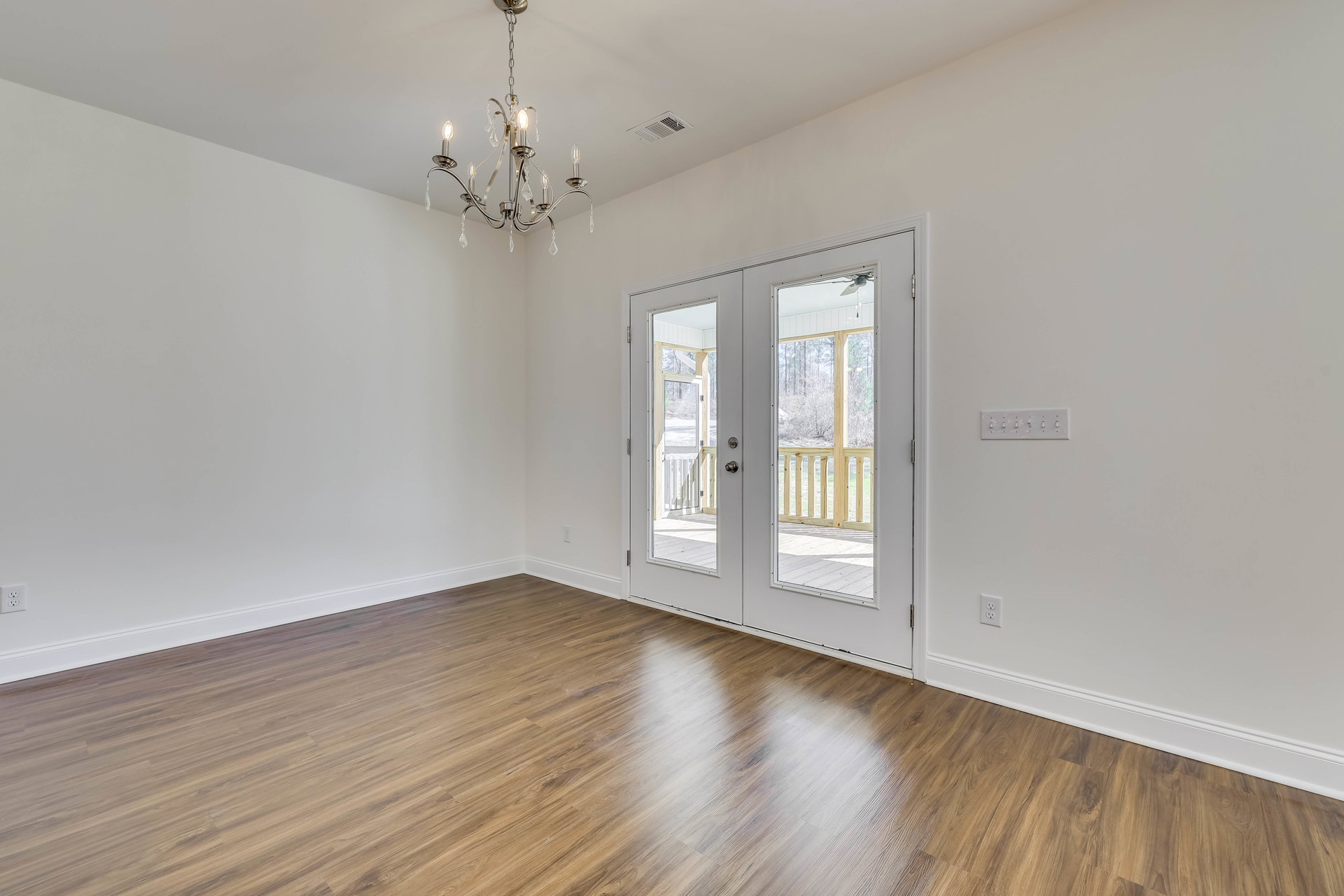 Chandelier hanging from ceiling above wood flooring, double glass doors open to outdoor view, white walls, large windows framing trees outside