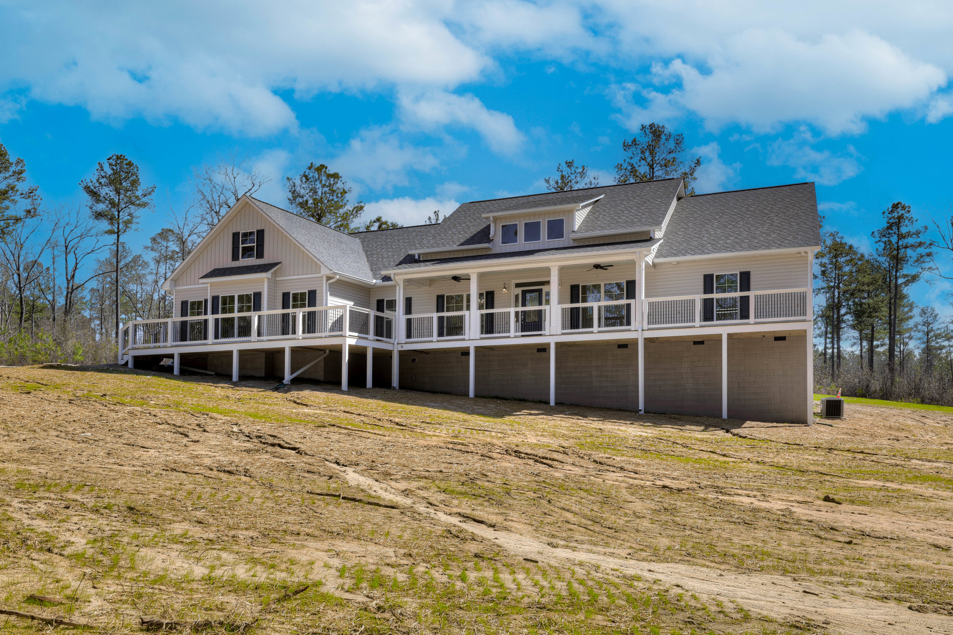 Two-story home with expansive wooden deck, light siding, multiple windows, sloped roof, surrounded by grass and mature trees