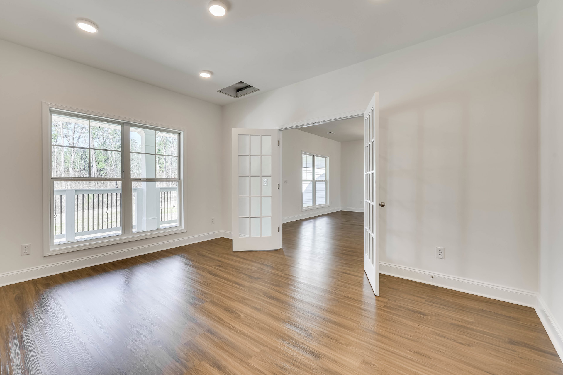 Wood floor room with open white door featuring glass panels, window with metal railing overlooking trees, plaster walls, ceiling vent