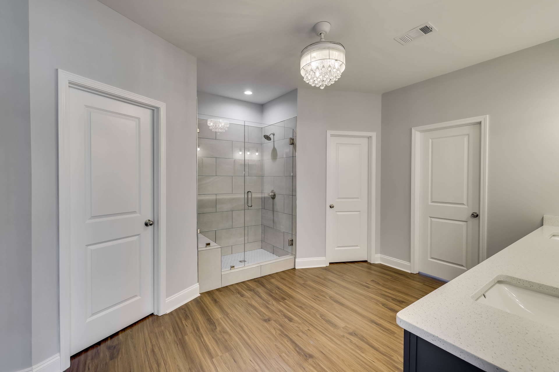 Bathroom with glass shower enclosure, wood plank flooring, white sink vanity, silver door knob on white door, clear glass chandelier, and white ceiling light fixture.