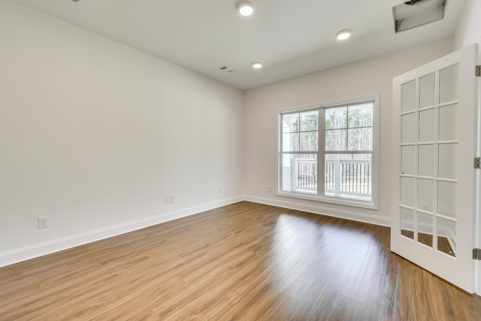 Wood flooring in a bright room with a large window, white door with glass panes, ceiling light, and view of trees and railing outside.