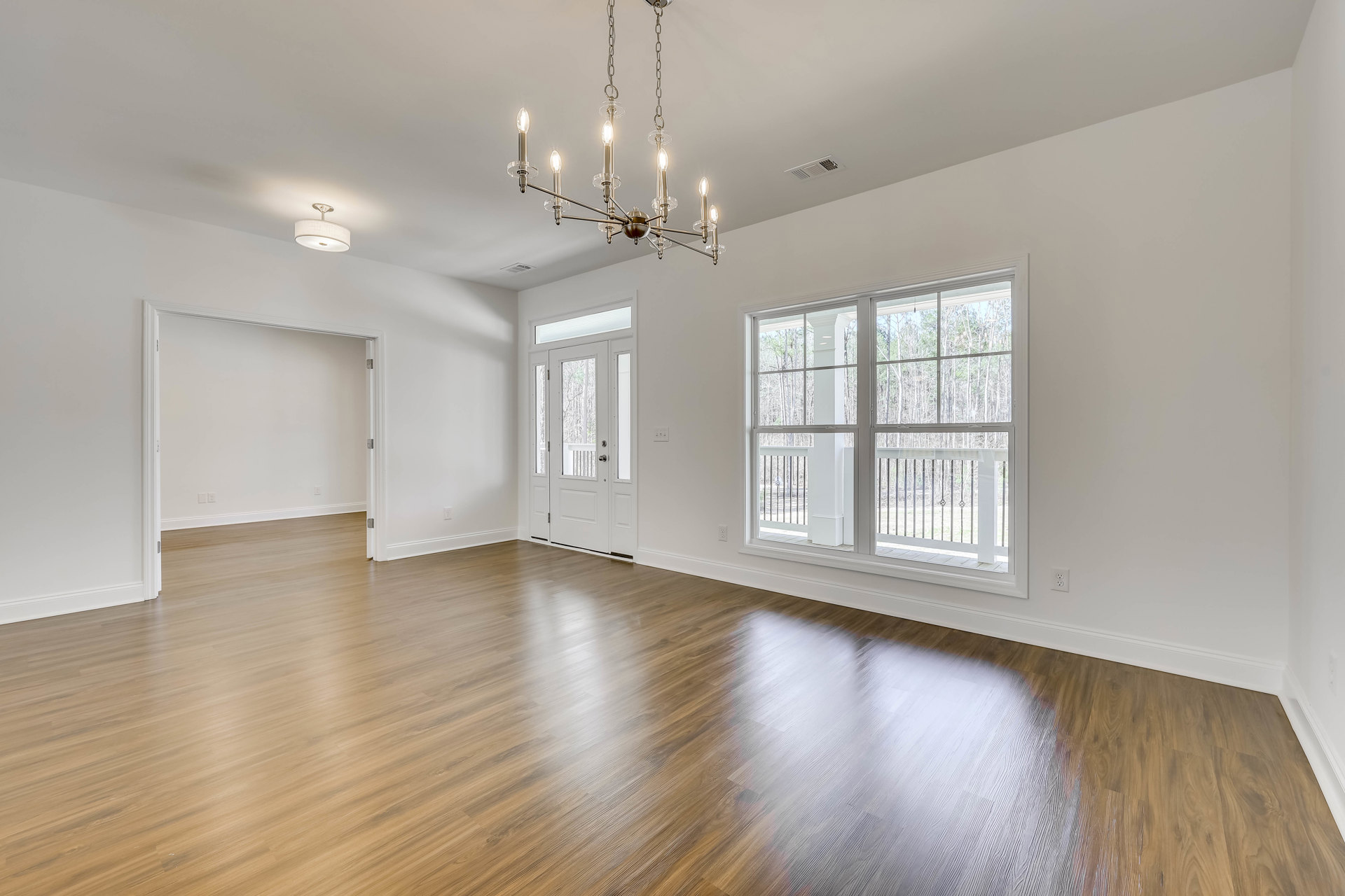 Chandelier hanging from ceiling above hardwood floor, white walls, large window with railing and view of trees, white door with glass panes