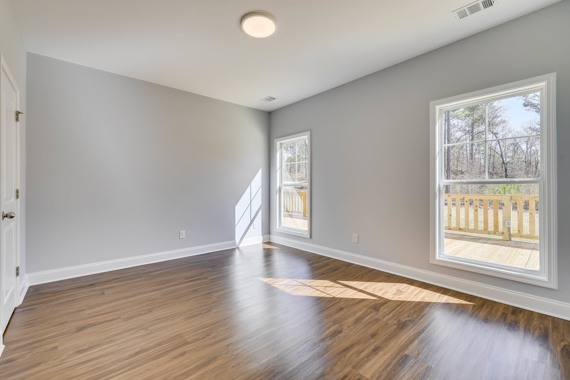 Room with light wood flooring, white plaster walls, large windows overlooking trees and deck, close-up of ceiling light fixture, wooden fence visible outside.