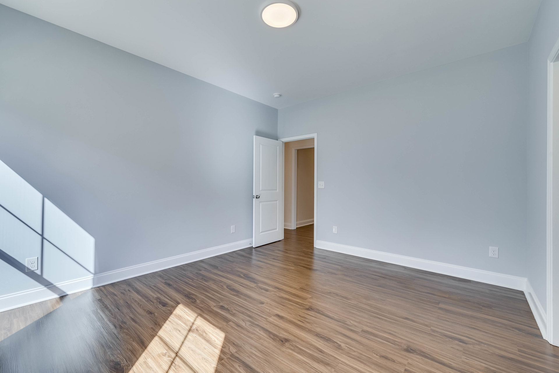 Wood flooring with white baseboard trim, open white door with silver knob, ceiling light fixture, smooth plaster walls