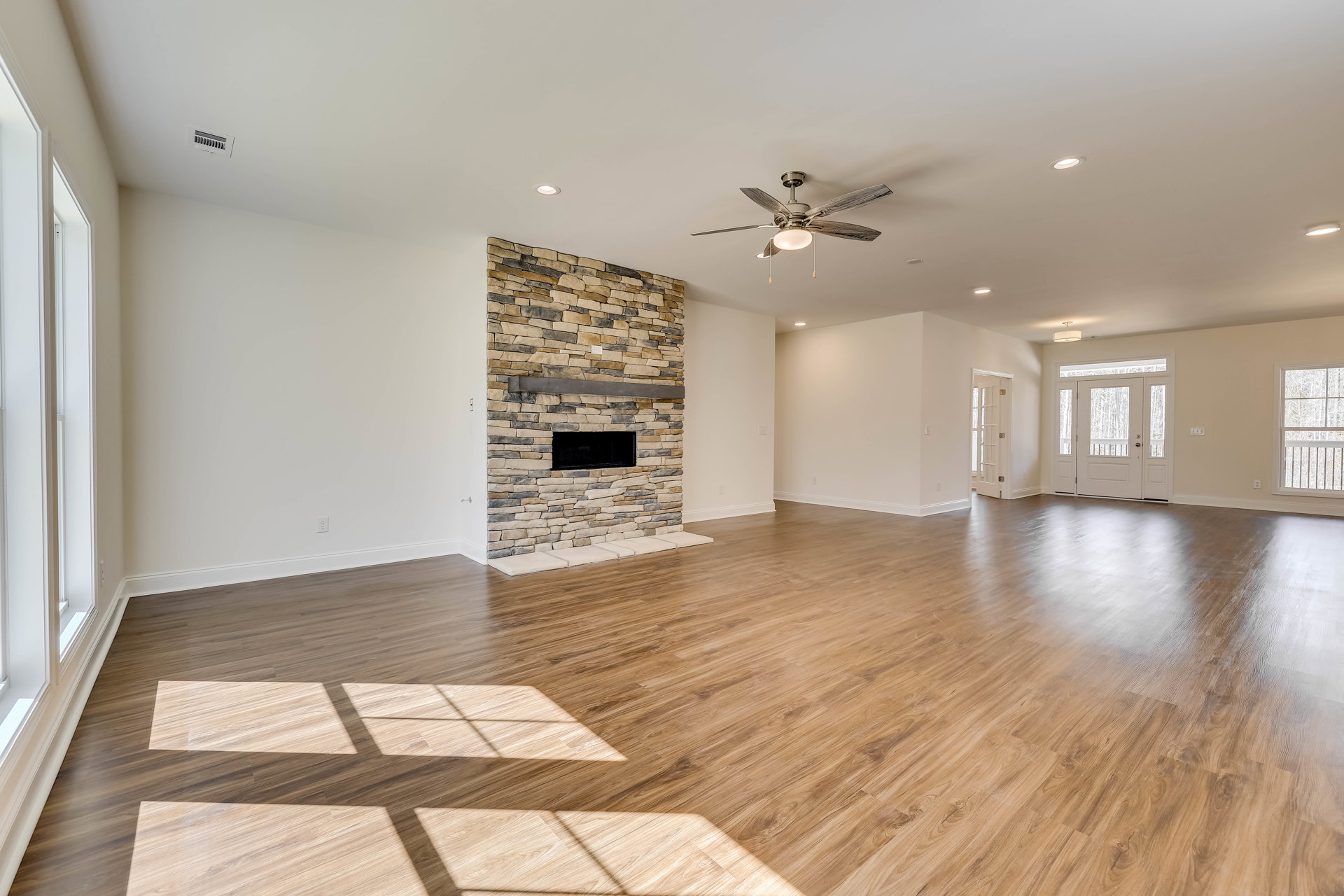 Spacious living room with wood flooring, stone fireplace featuring a black firebox, ceiling fan with light, plaster walls, and large window overlooking trees and railing.
