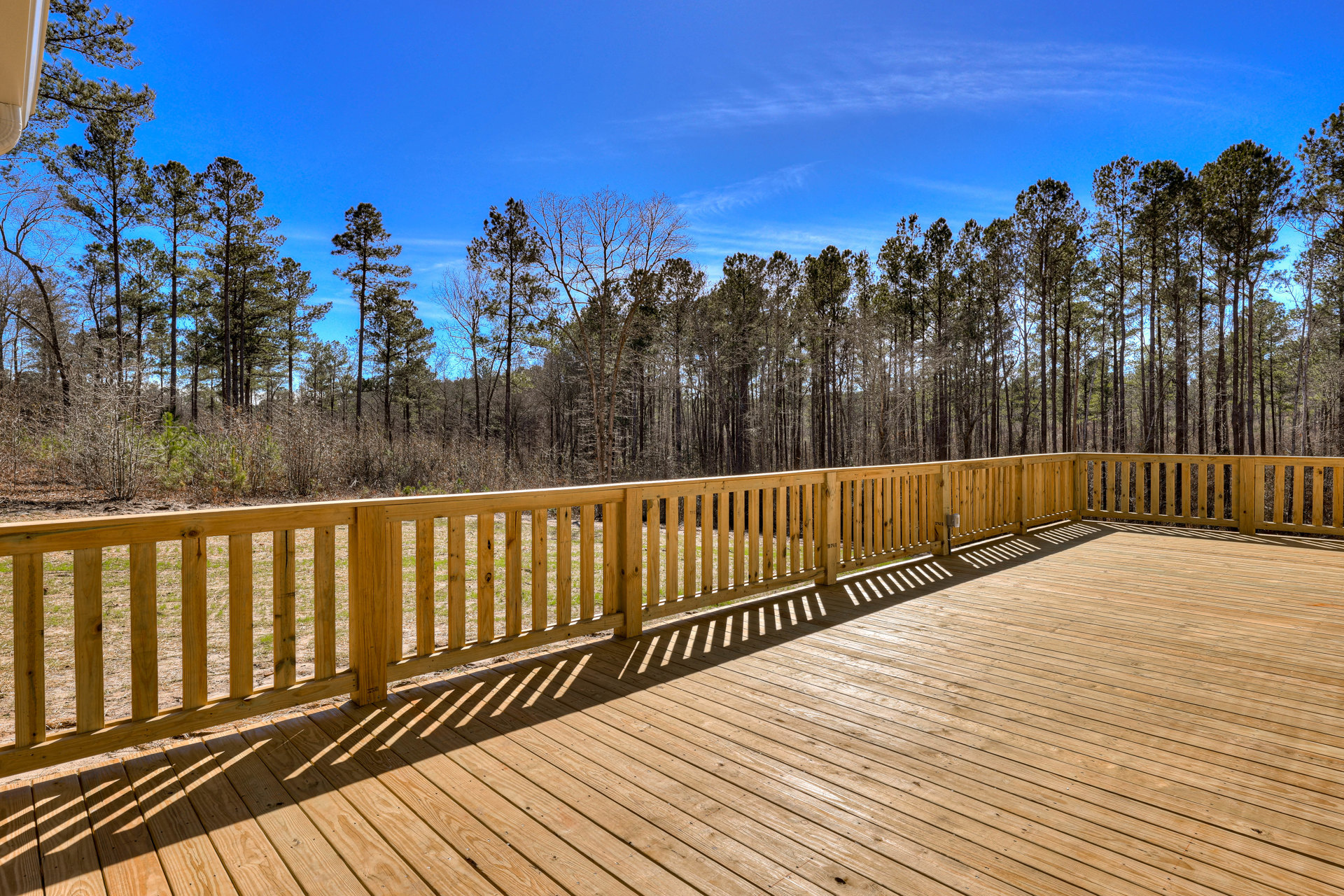 Wooden deck with horizontal wood railing, surrounded by tall trees and a partly cloudy blue sky in the background