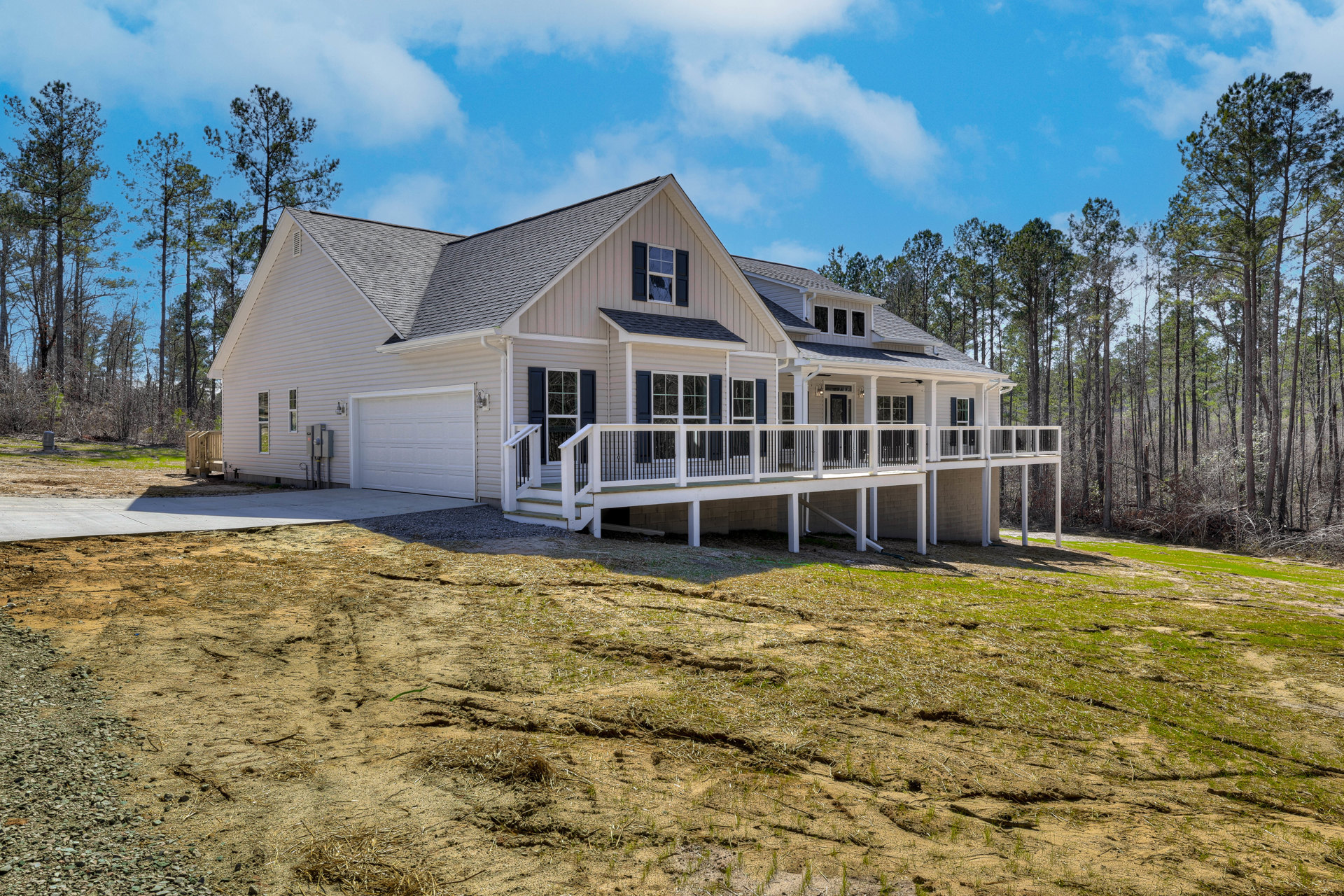 White two-story home with expansive white deck featuring black railing, white-framed windows, brick accent wall near garage door, dirt hill in foreground, and blue sky overhead
