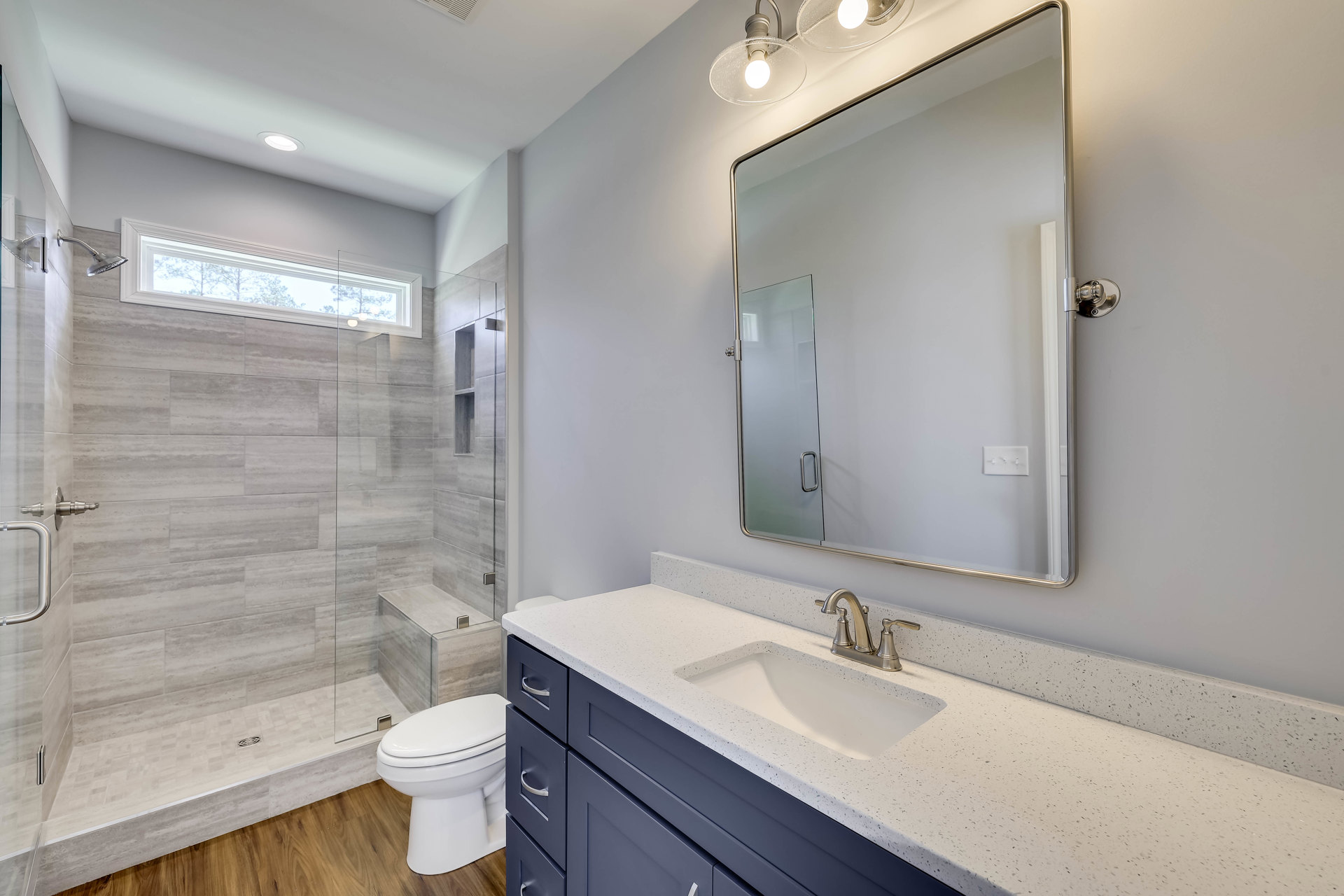 Modern bathroom with white tile walls, glass shower enclosure, rectangular mirror above a stone countertop sink, chrome faucet, and white toilet
