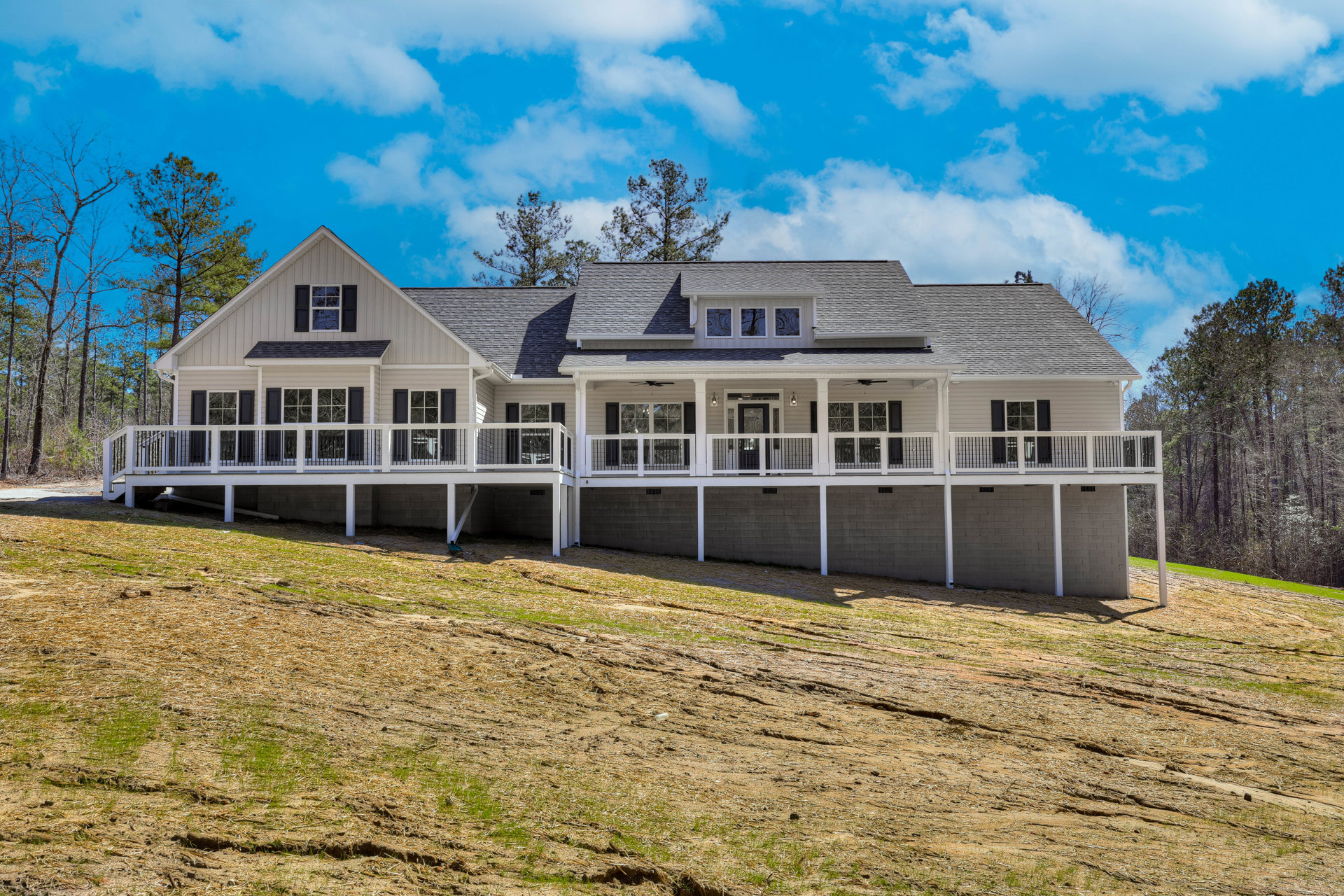 Spacious front porch with white railings, light gray siding, large windows, gabled roof, and surrounding green lawn with mature trees