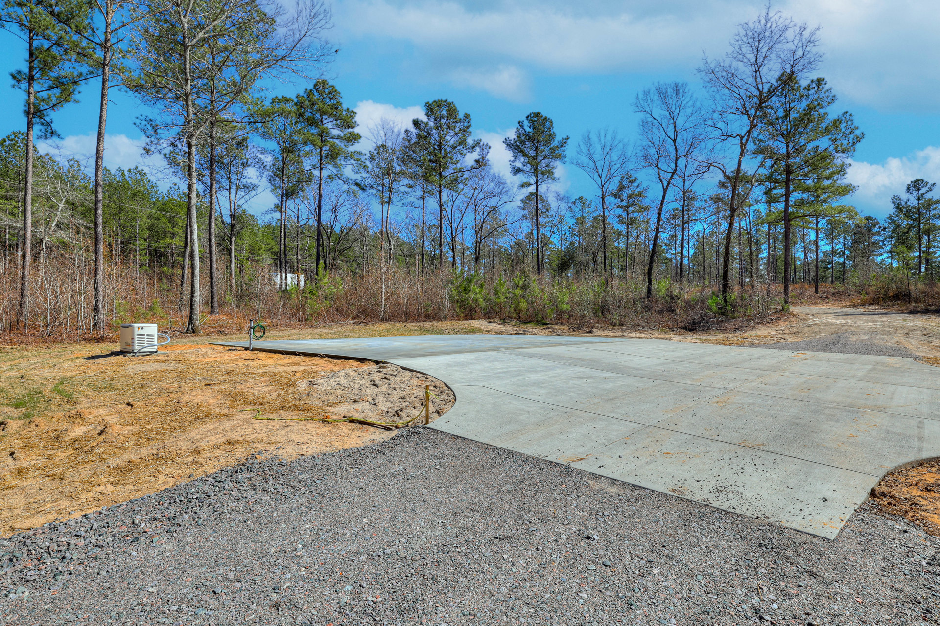 Gravel driveway bordered by grass and dense trees, white generator positioned on ground, French Creek State Park forest visible in background under cloudy sky